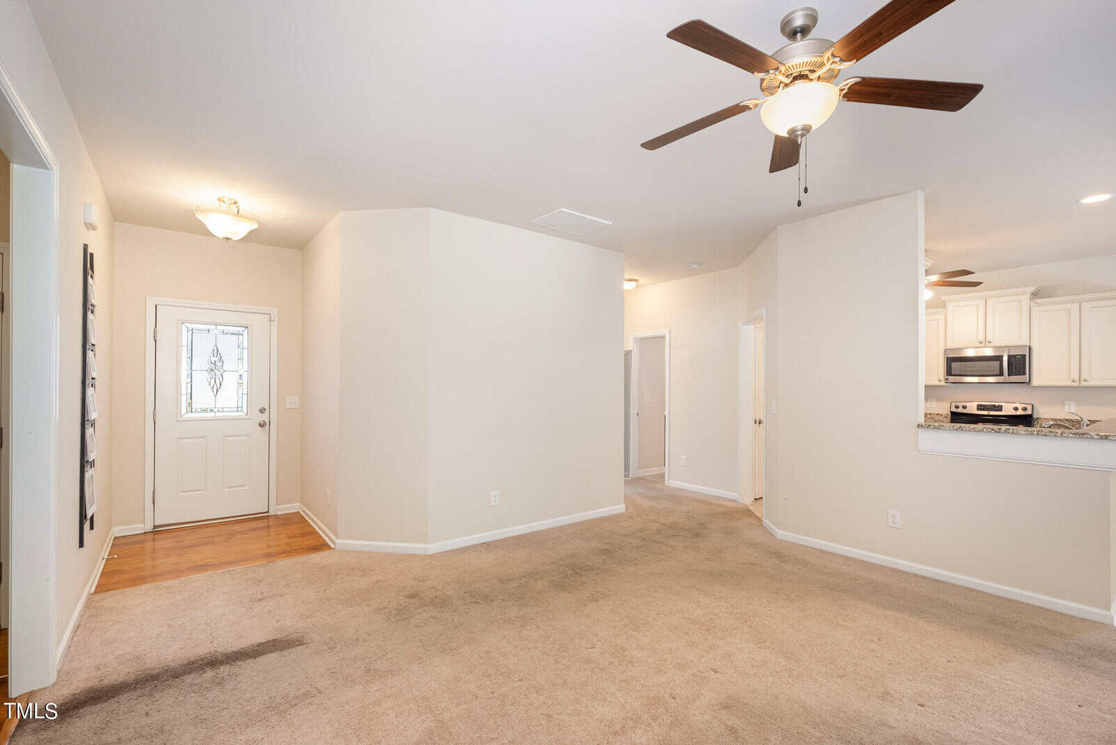A spacious, empty room with beige carpet and light-colored walls. It features a ceiling fan with lights, a kitchen area with stainless steel appliances in the background, and a white front door. Natural light illuminates the space.