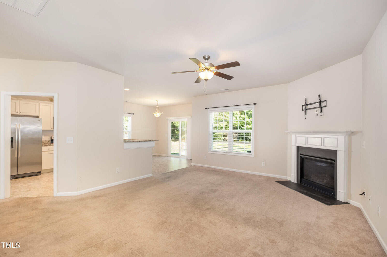 Spacious living room with beige carpet, white walls, and a ceiling fan. A fireplace is in the corner, and large windows let in natural light. The room leads to a bright kitchen with stainless steel appliances and a dining area.