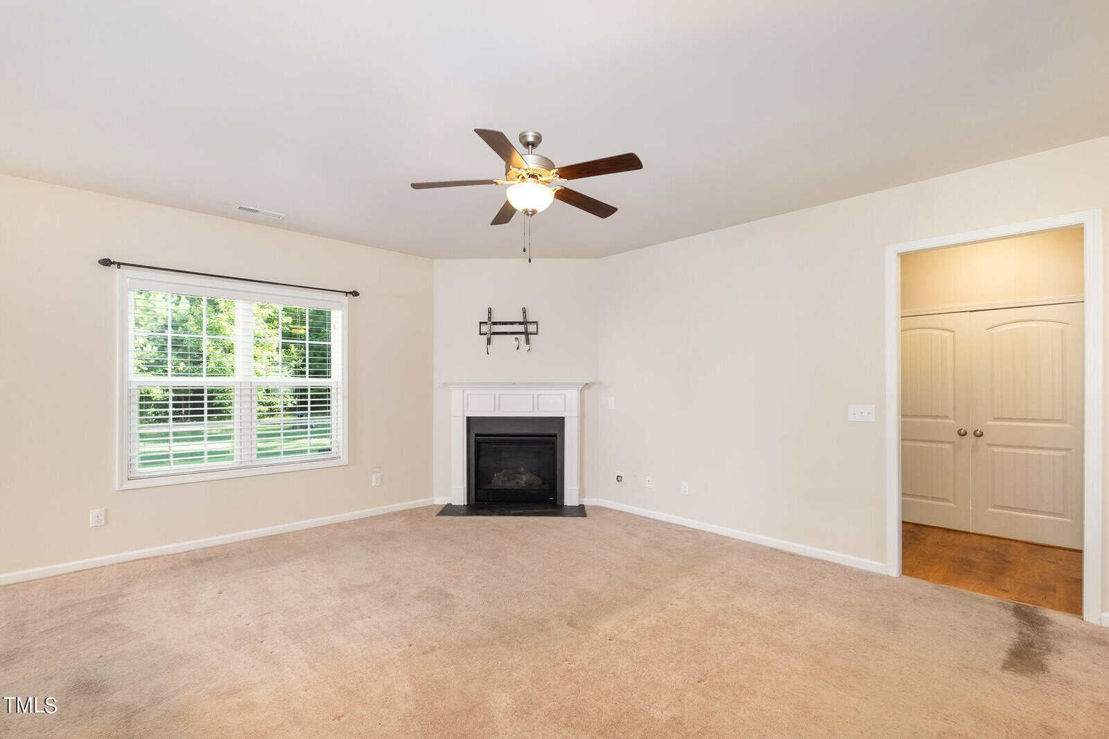 A spacious, empty living room with beige walls and carpet, featuring a white fireplace with a TV shelf above. A ceiling fan with lights hangs from the ceiling. Large window with blinds on the left lets in natural light. Doorway visible on the right.