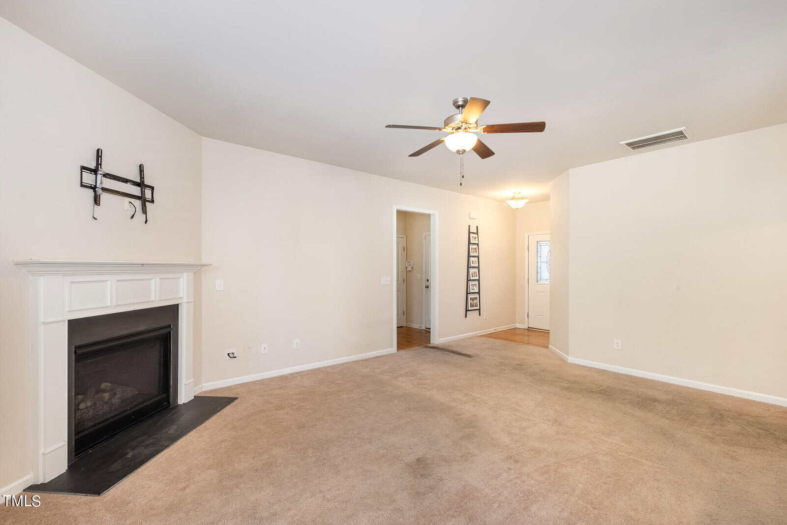 Spacious empty room with beige carpet, white walls, and a ceiling fan. A fireplace with a mantle is on the left, and multiple framed photos are displayed on the wall near an open doorway on the right.
