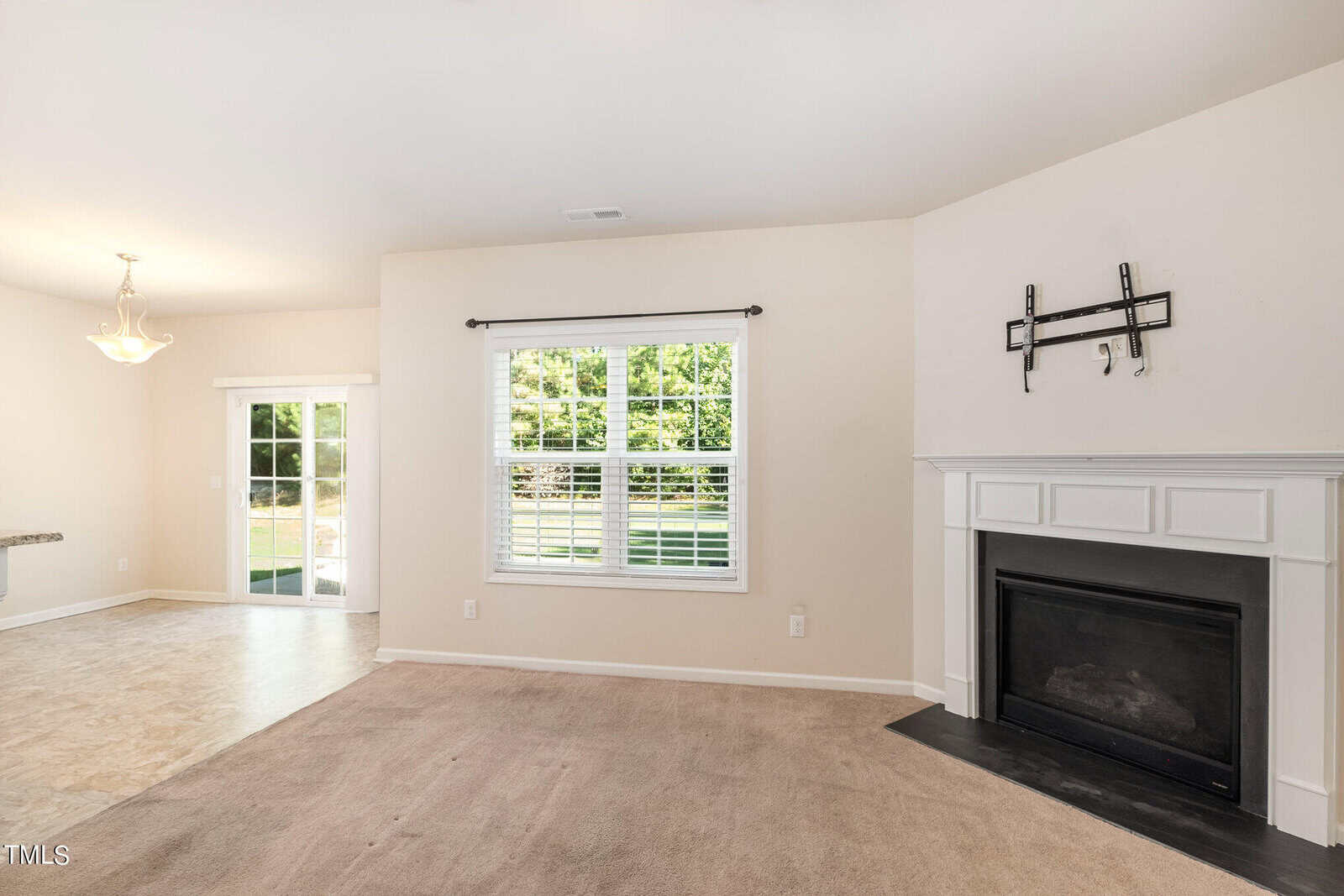 Living room with beige carpet, a white fireplace, and wall mount for a TV. Large window with blinds overlooks a grassy yard. Adjacent dining area with tiled floor and hanging light fixture. Sliding glass door leads to an outdoor space.