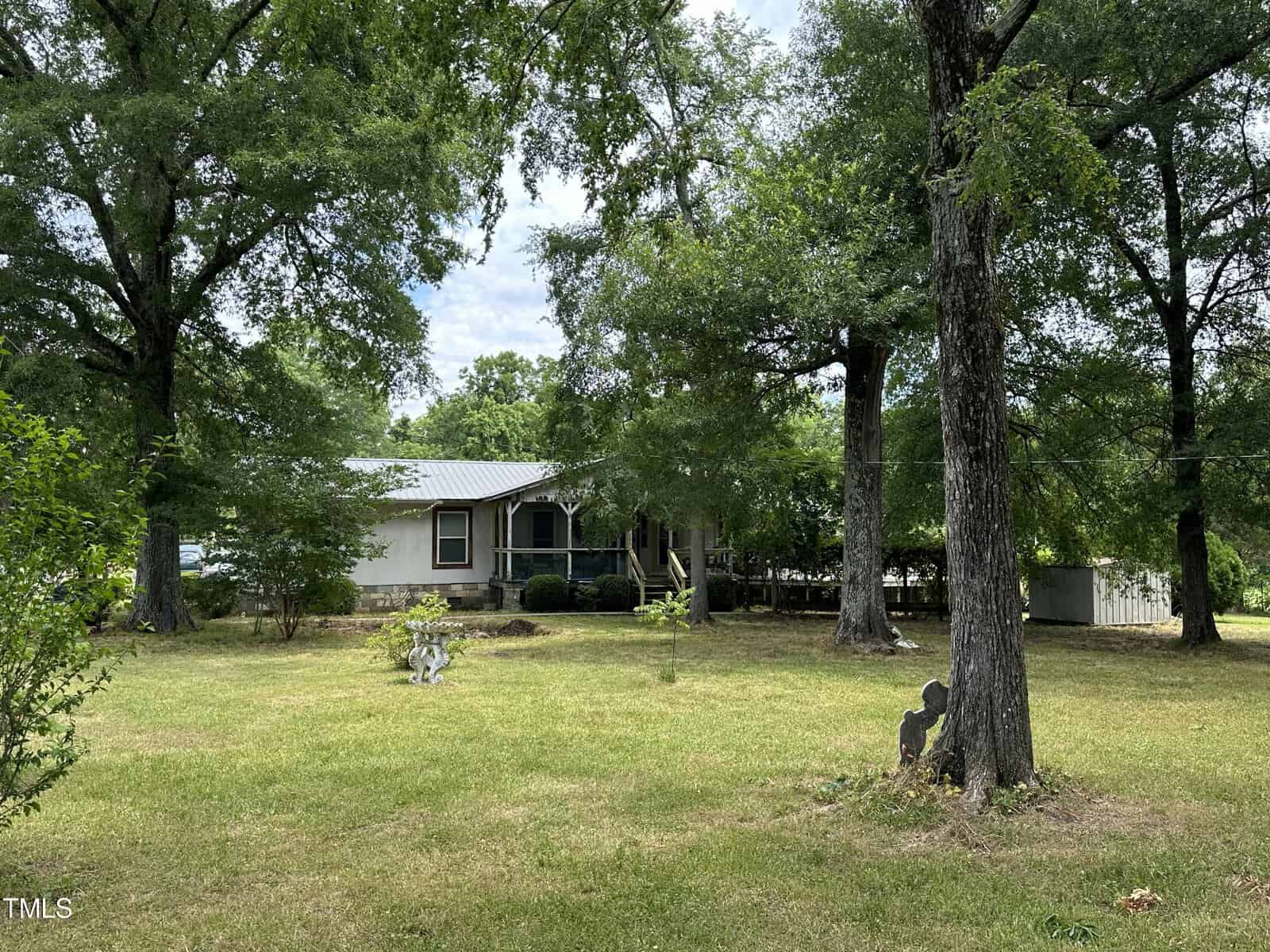 A rural house with a metal roof is surrounded by large trees and a grassy yard. There is a porch with railings at the front and a wooden shed in the background. A silhouette of a child and dog statue is visible in the yard.