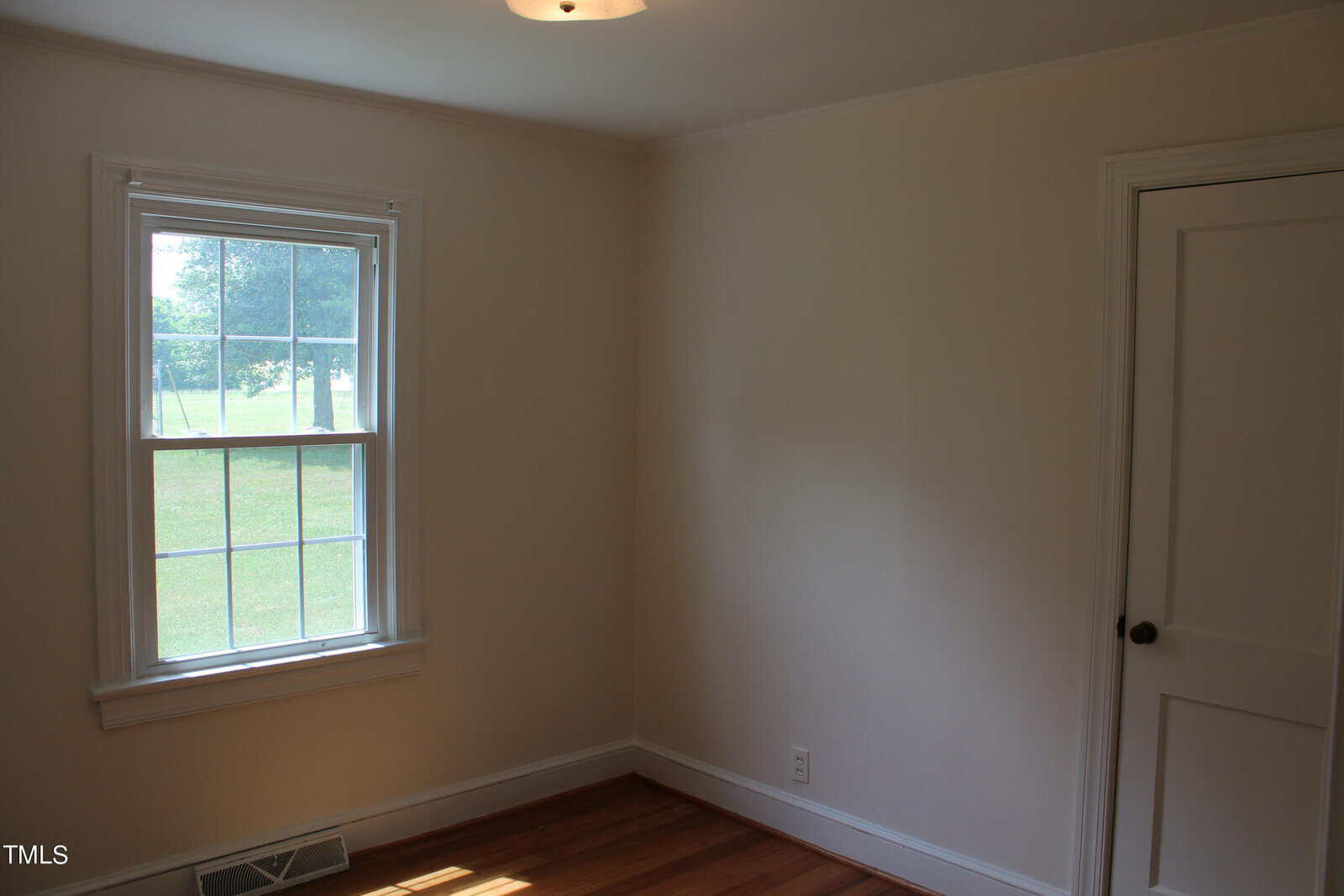 A small, empty room with white walls and wooden flooring. A single window on the left allows natural light to enter, revealing a view of green grass and trees outside. A closed white door is on the right. A ceiling light fixture is visible.