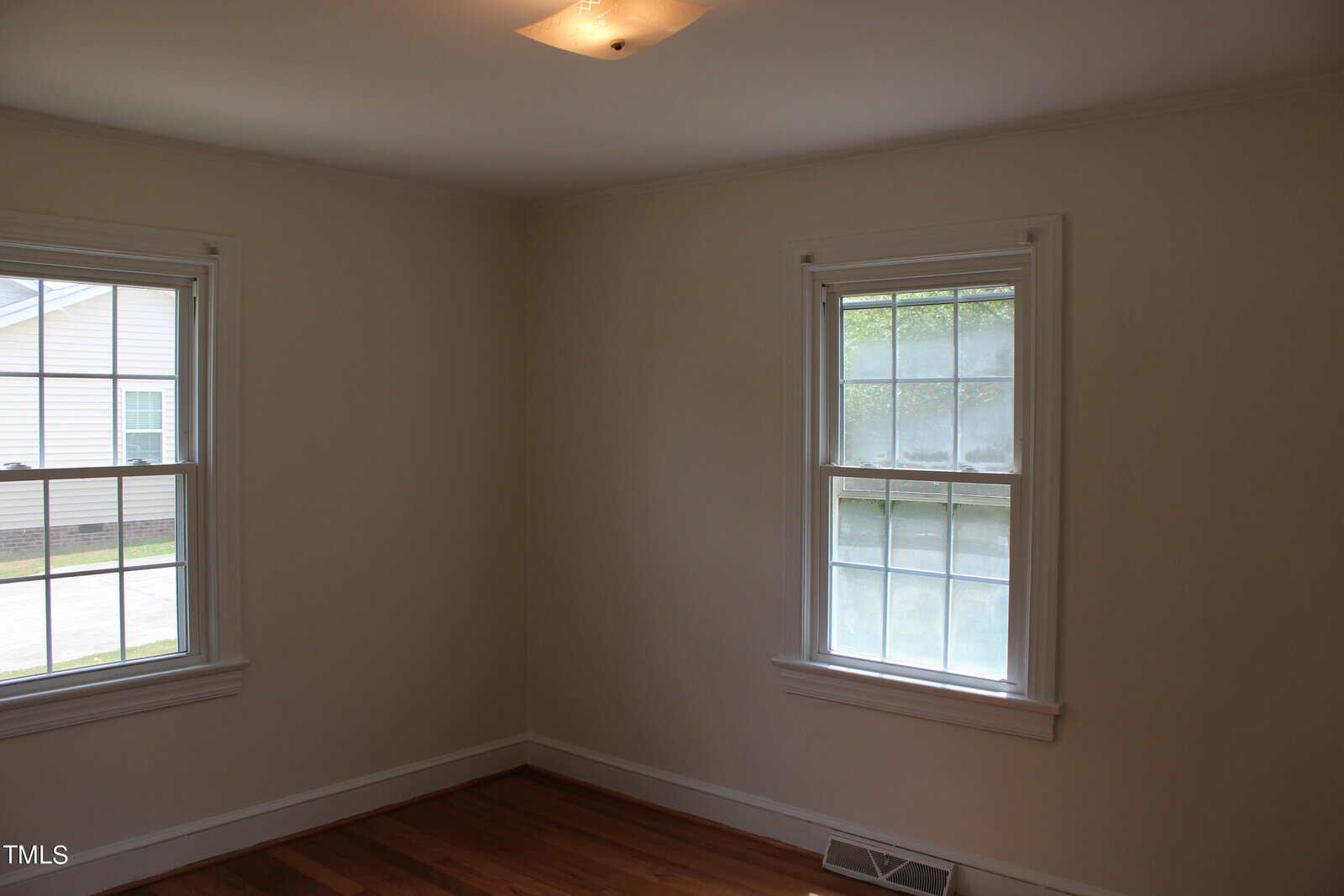 A small, empty room with white walls and hardwood floors. Two windows allow natural light to brighten the space. A simple ceiling light fixture is visible. The view outside shows parts of a neighboring building.