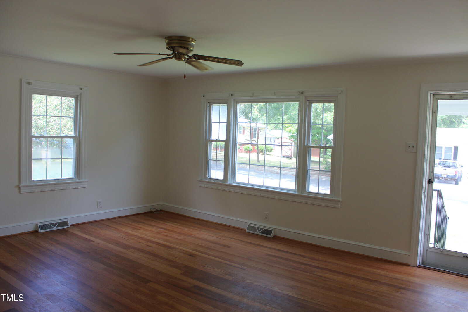 An empty room with wooden floors, white walls, and two large windows with views of trees and a house. A ceiling fan is centered above, and there's a door leading outside. The room is well-lit with natural light.