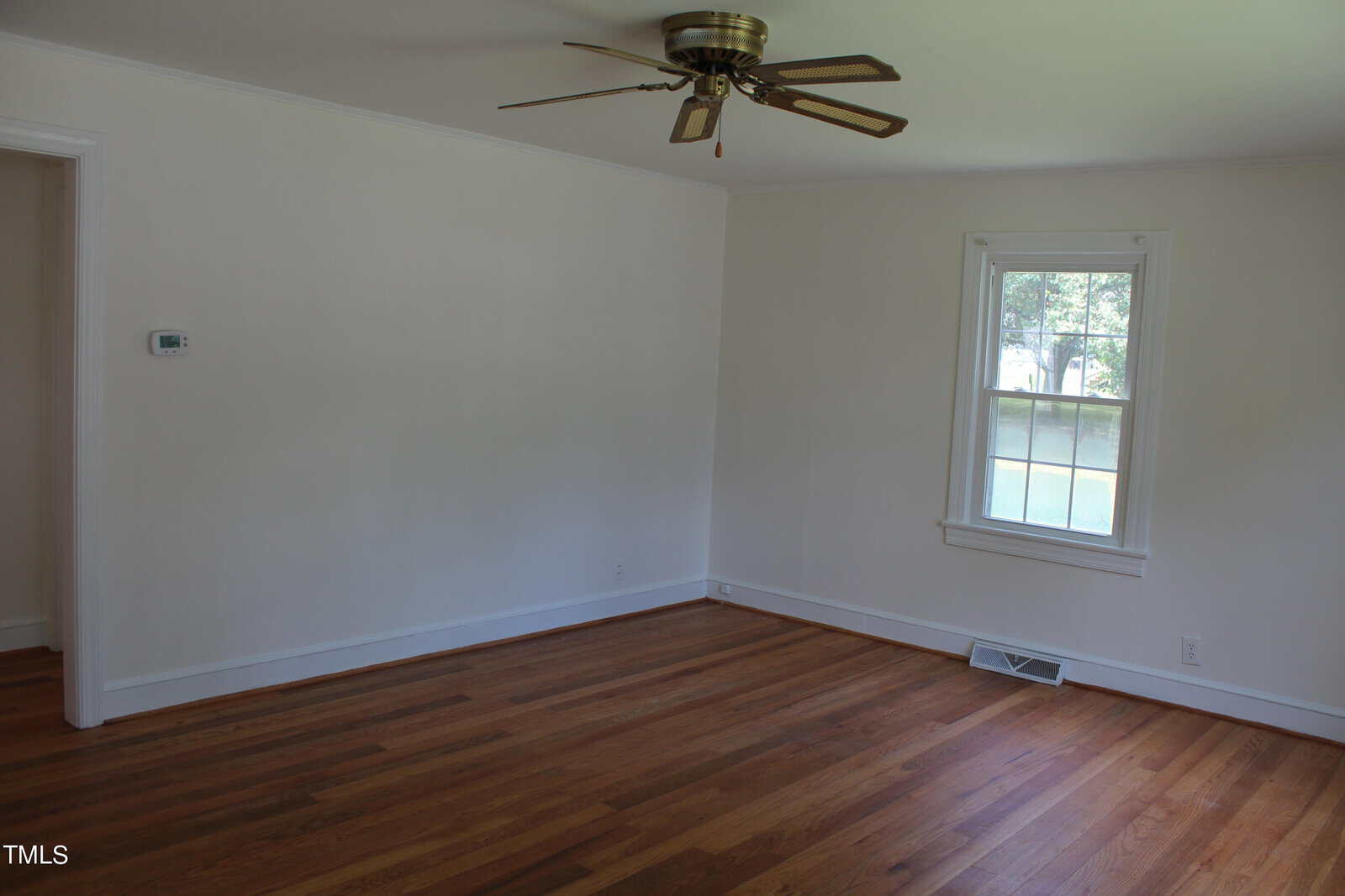 An empty room with wooden floors, white walls, a ceiling fan, and a single window letting in natural light. A vent is located below the window.