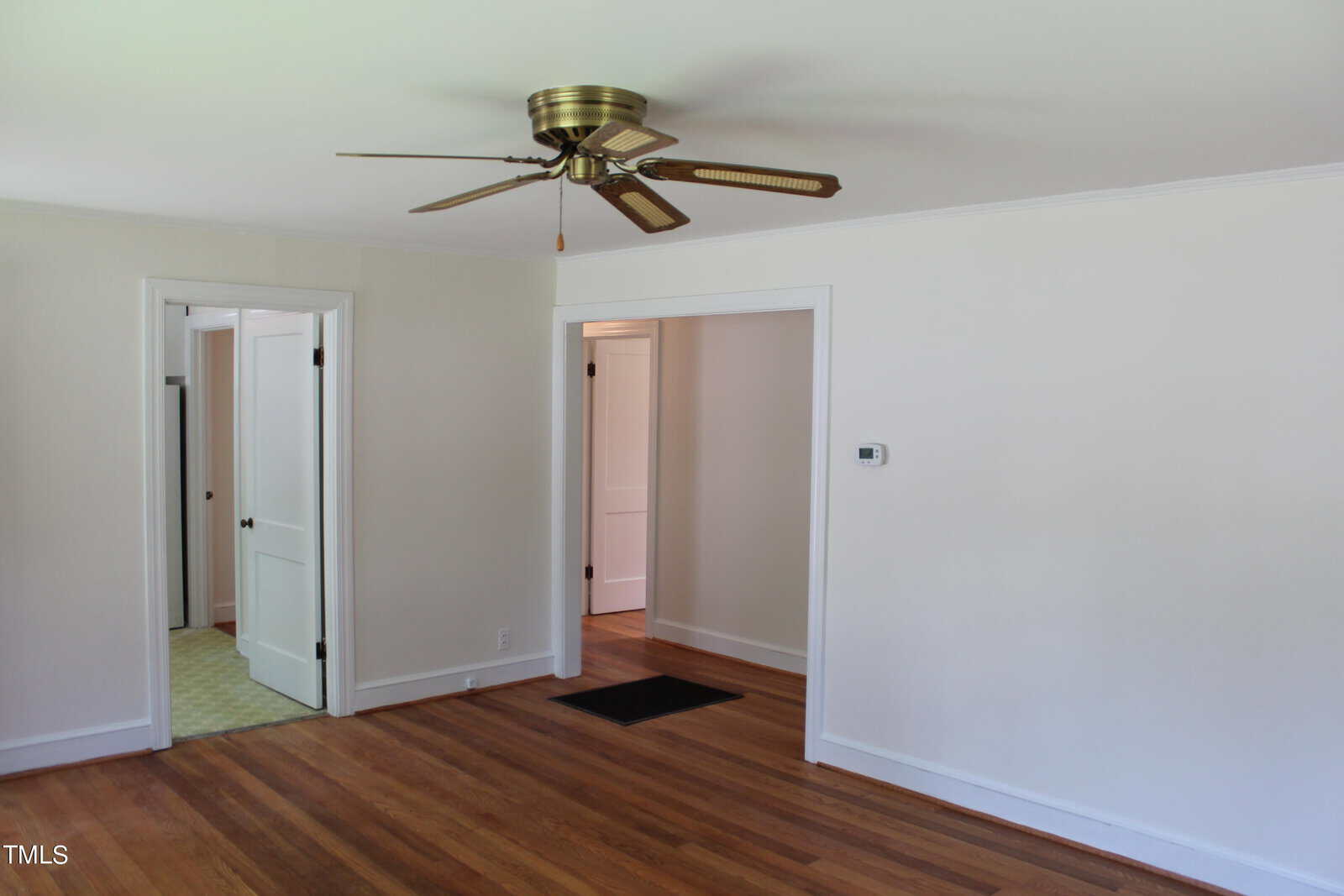 An empty room with light-colored walls and wooden flooring. There's a ceiling fan centered on the ceiling, and two open doorways are visible, leading to adjacent rooms.