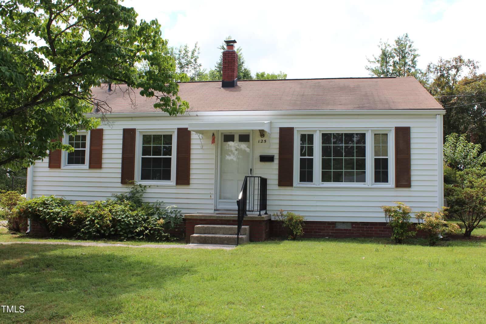 The small white house at 125 Kearney Avenue in Oxford features charming brown shutters, a red brick chimney, and a well-kept front yard. Two large windows frame the exterior, with steps leading up to the inviting white front door. Trees and bushes provide a lush backdrop to this picturesque property.