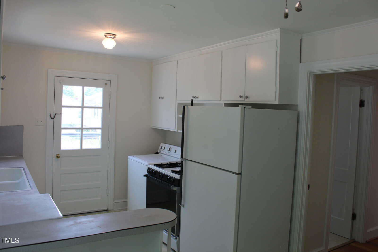 A small kitchen with white cabinets, a white refrigerator, a gas stove, and a sink. There's a windowed door at the end of the room allowing natural light. The walls are painted white, enhancing the brightness of the space.