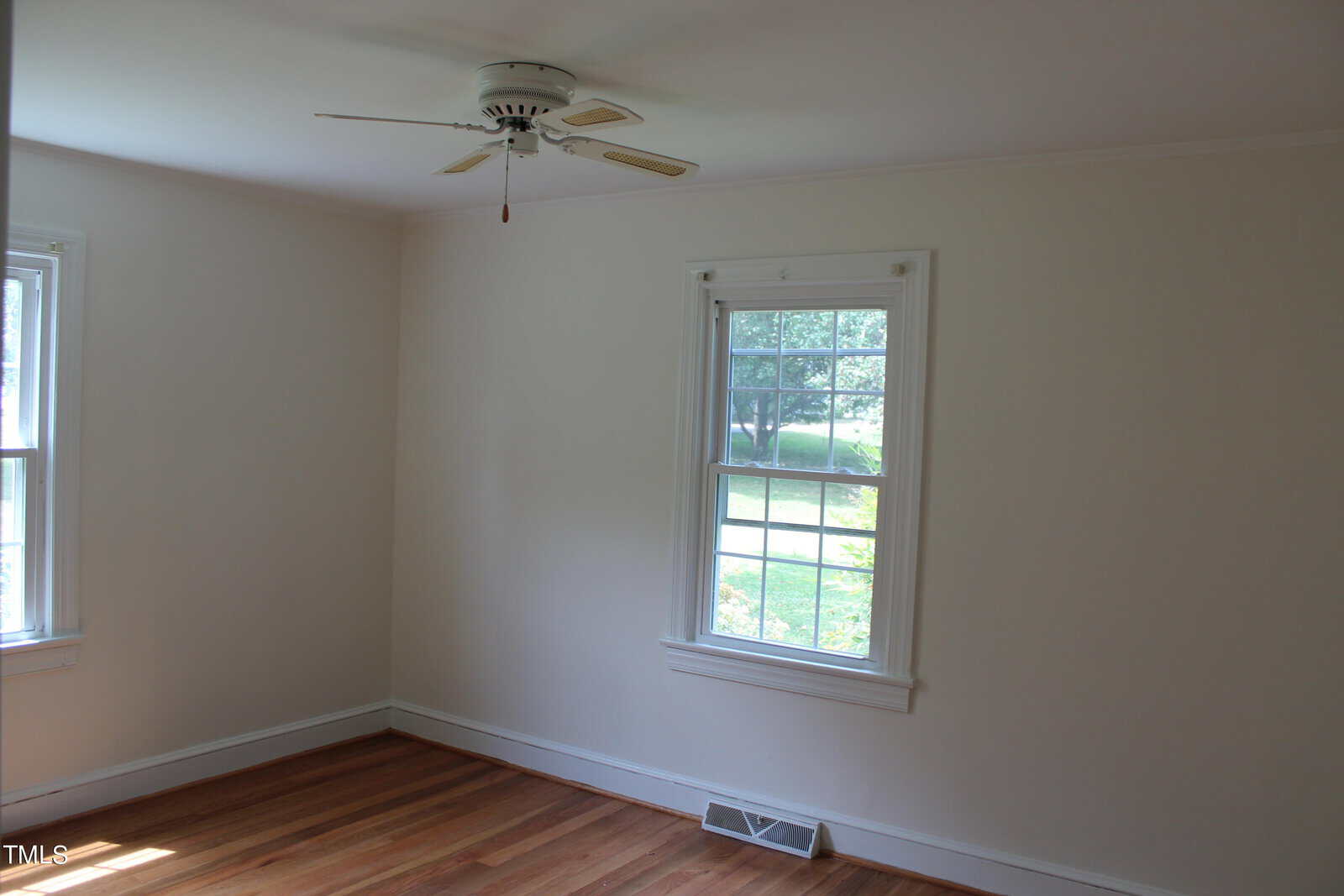An empty room with white walls, a ceiling fan, two windows showing a green outdoor view, and hardwood floors. There's a wall vent below one window.