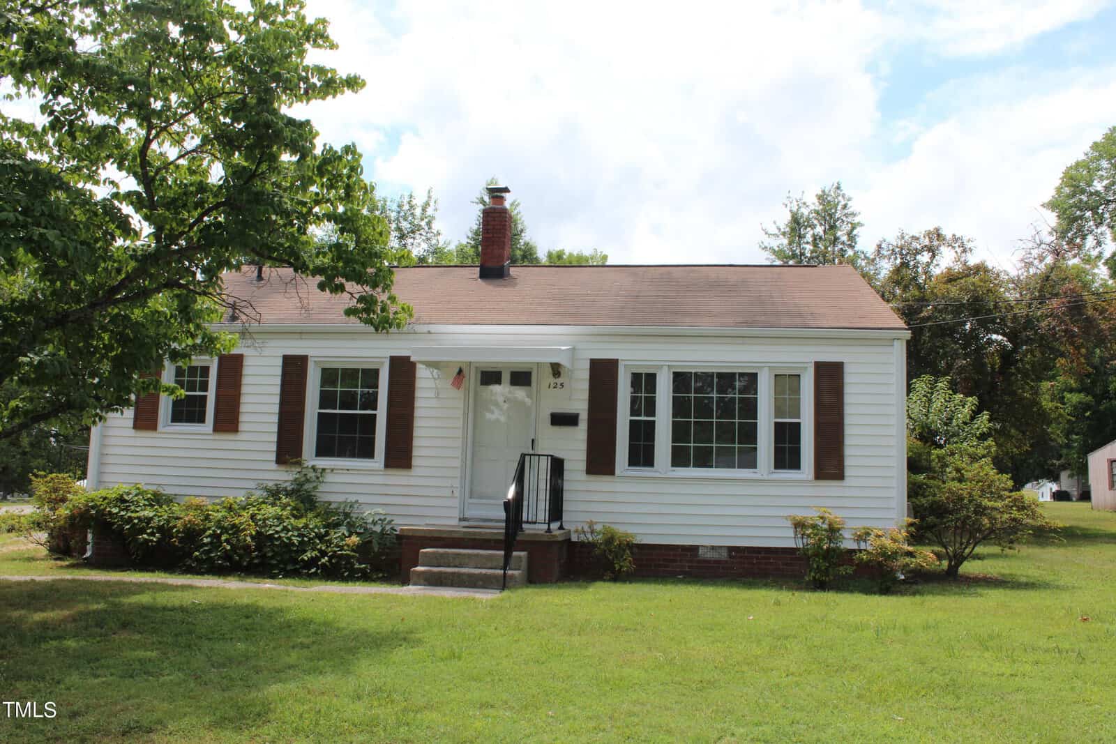 A small, single-story white house with brown shutters and a brick chimney, surrounded by green lawn and trees. Steps lead up to a black door with a small porch. The sky is partly cloudy.