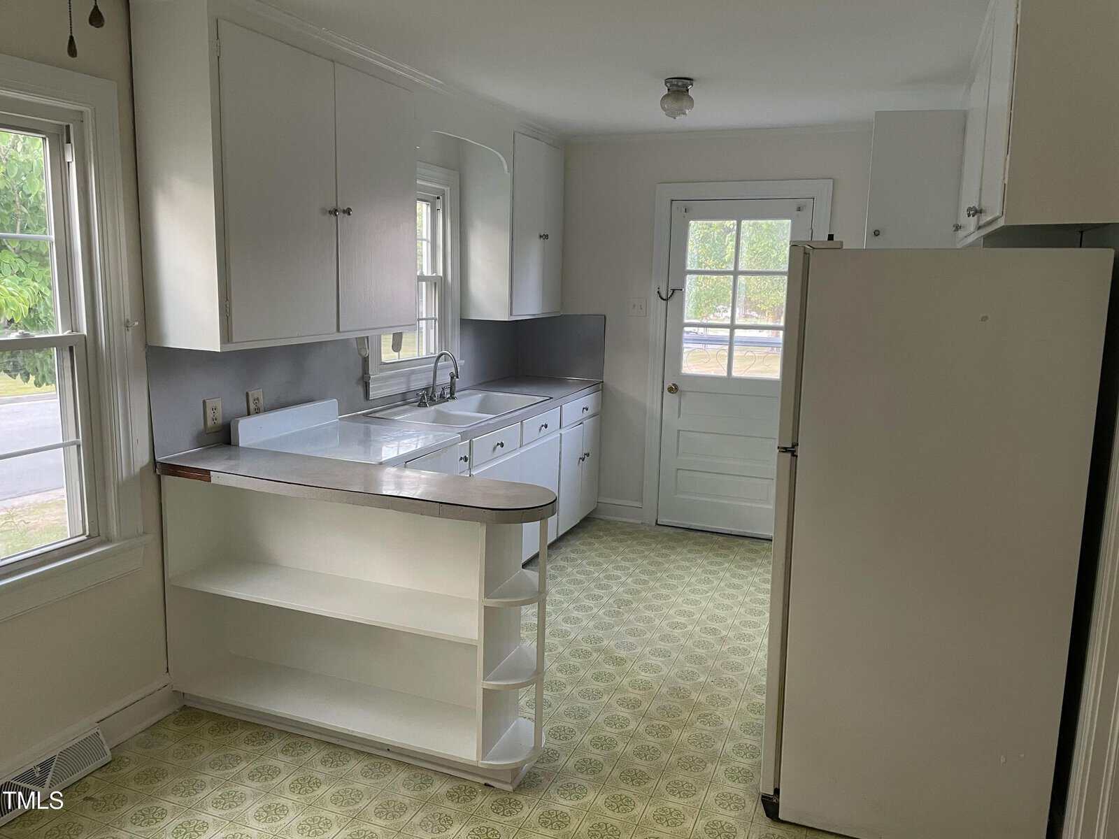 A bright, vintage kitchen with white cabinets and countertops. There's a double sink under a window, a refrigerator on the right, and a door leading outside. The room features yellow patterned flooring and a small open shelf near the sink.