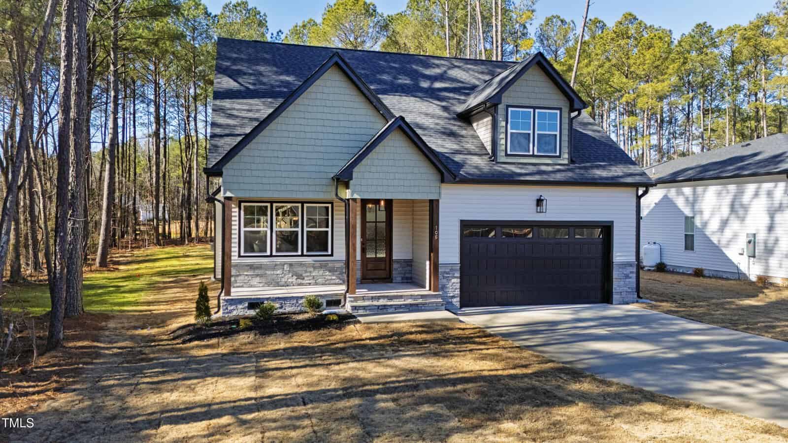 A modern two-story house with light green siding, stone accents, and a dark roof, featuring a covered front porch, large windows, and a two-car garage. The house is surrounded by tall trees and sits on a spacious lot.