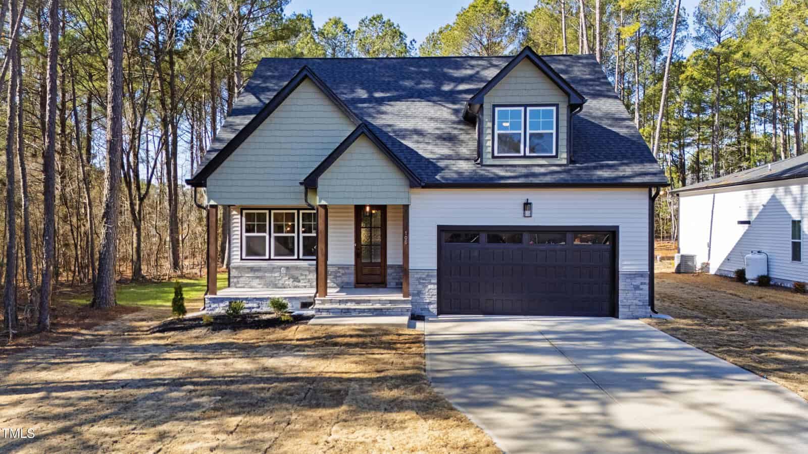 A modern two-story house with green siding, dark roof, and attached double garage, set among tall trees with a concrete driveway and a small front porch. The yard is mostly brown grass with some landscaping.