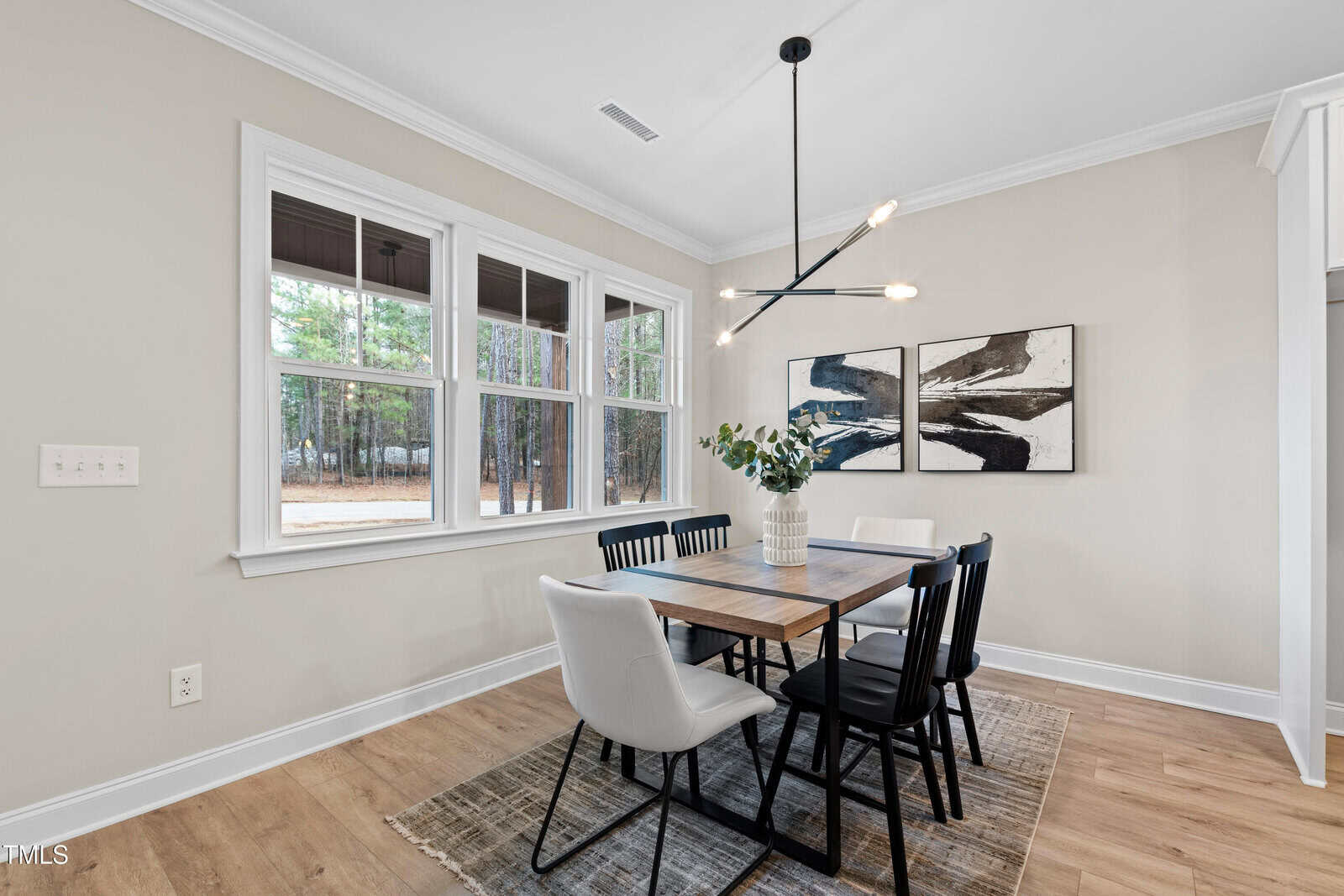 A modern dining area with a wooden table, black and white chairs, a contemporary chandelier, a vase with green branches, abstract wall art, large windows, and light wood flooring.