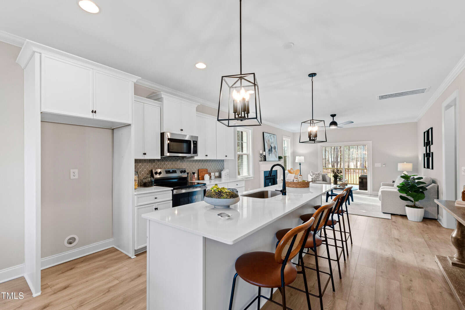 Modern kitchen with white cabinets, stainless steel appliances, a large island with sink, three barstools, pendant lighting, and an open layout leading to a bright living area with large windows and wood flooring.