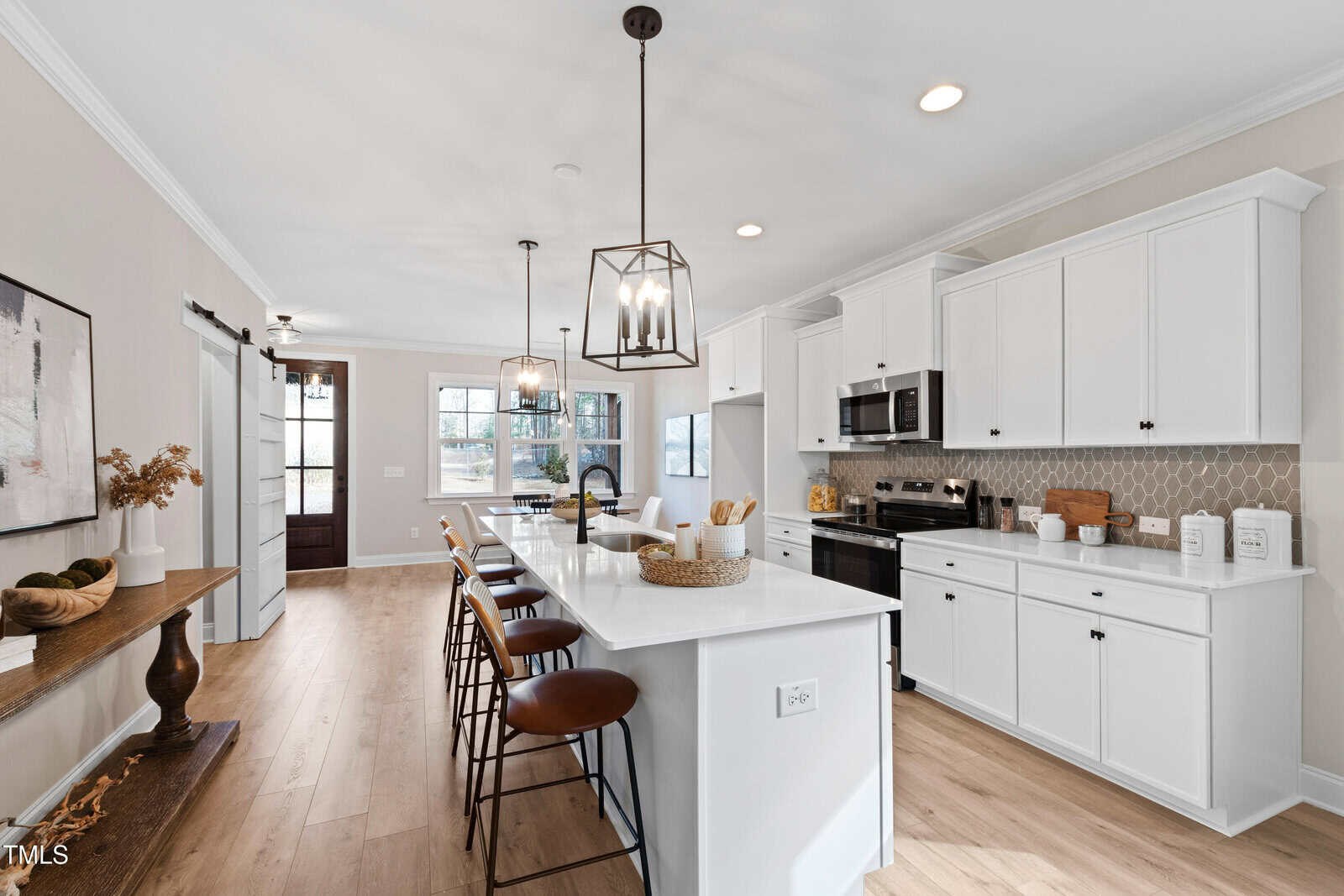 Modern kitchen with white cabinets, a large island with bar stools, hanging pendant lights, light wood flooring, stainless steel appliances, and a decorative shelf with plants and vases along one wall.