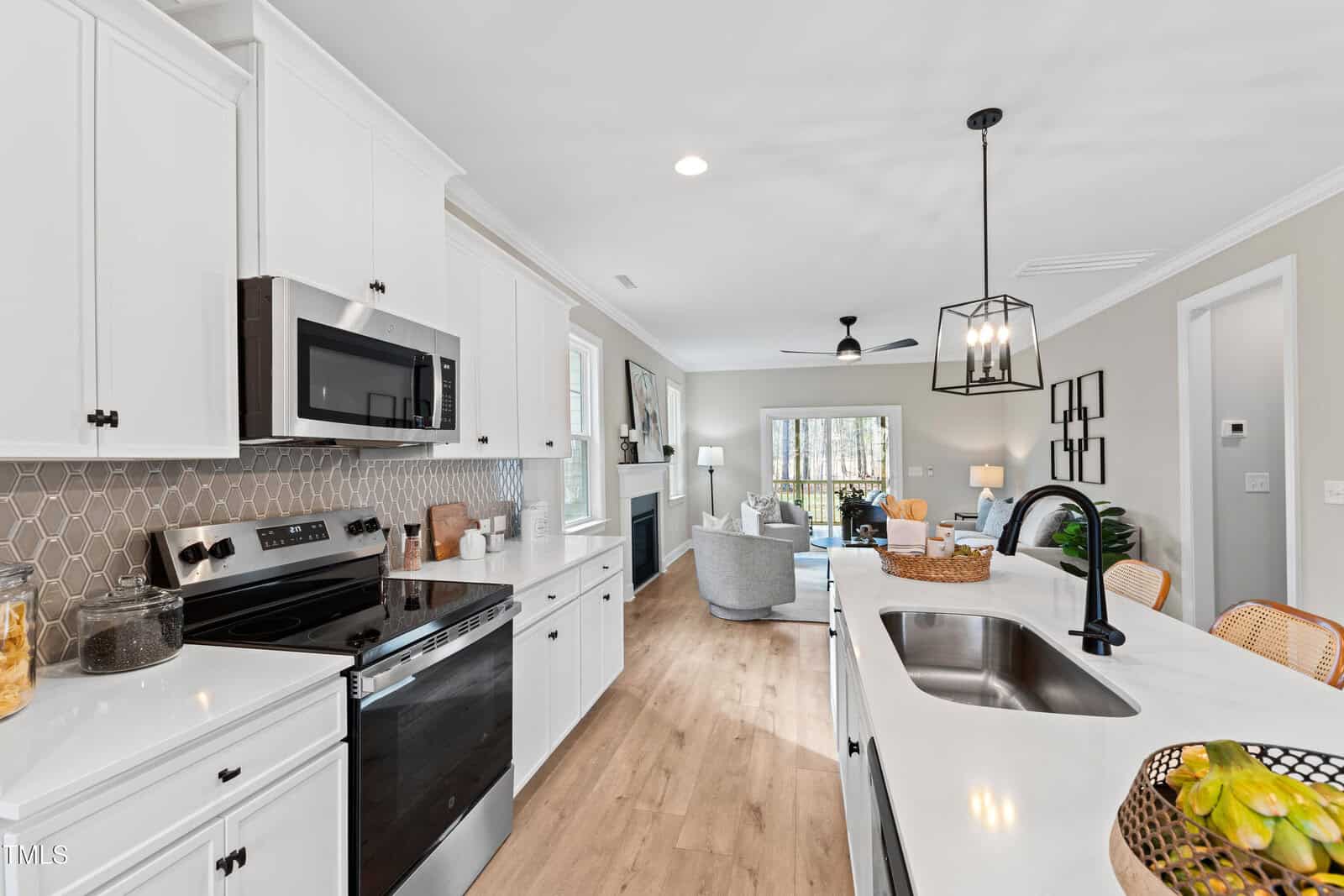 Modern kitchen with white cabinets, black appliances, and a hexagon tile backsplash, opening into a bright living area with a fireplace, cozy seating, and large windows. A black pendant light hangs over the kitchen island.