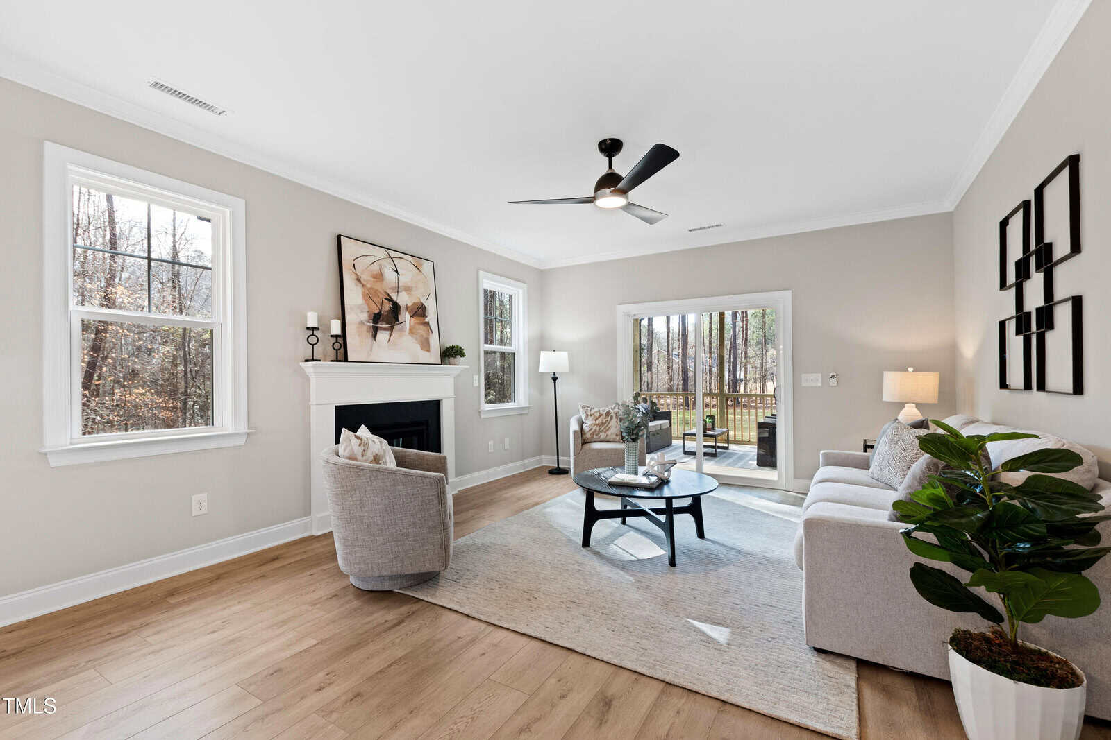 Bright living room with light wood floors, a gray sofa, two gray chairs, a round coffee table, and a fireplace. Large windows and glass doors offer views of trees outside, filling the space with natural light.
