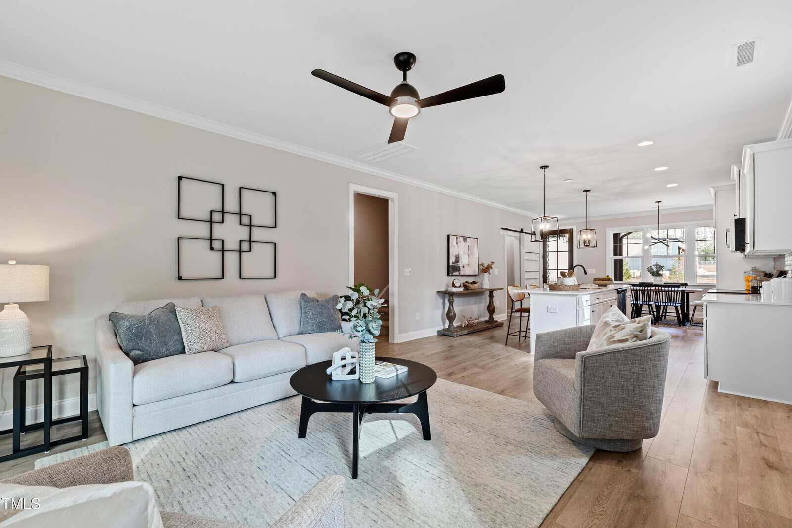 A modern open-concept living room and kitchen with light wood floors, neutral furniture, a round coffee table, wall art, a ceiling fan, and large windows letting in natural light.