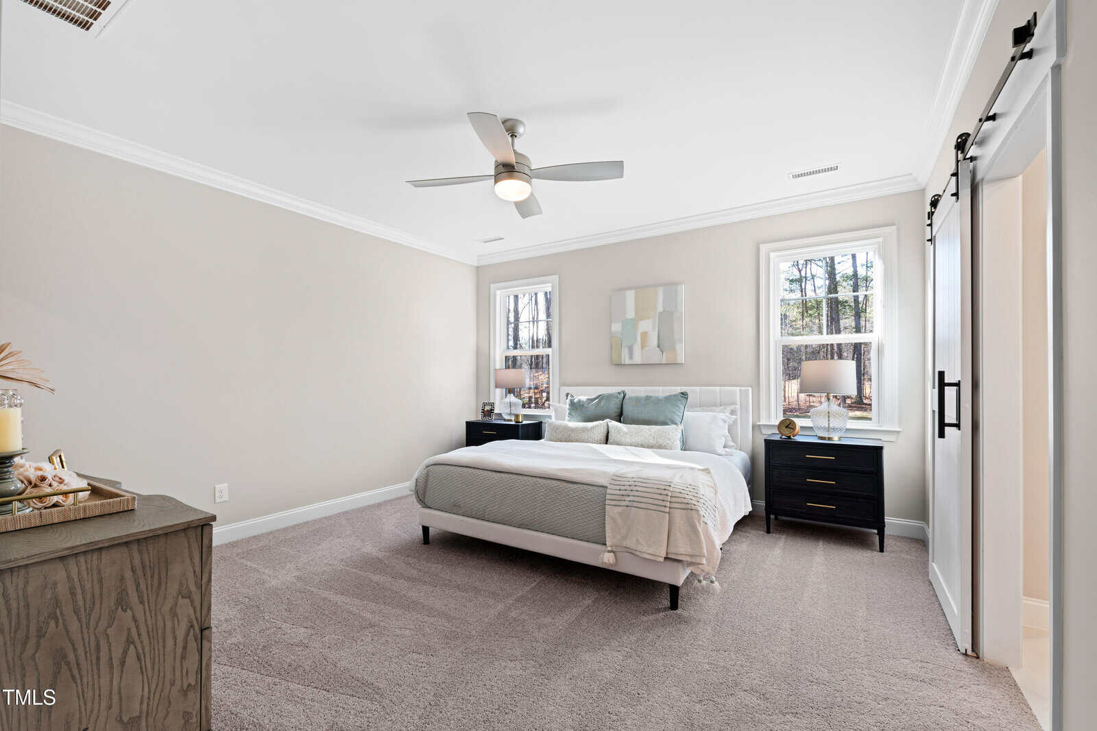 A bright bedroom with light beige walls, large windows, a bed with white and pastel bedding, two black nightstands, a ceiling fan, carpeted floor, and a sliding barn door on the right.