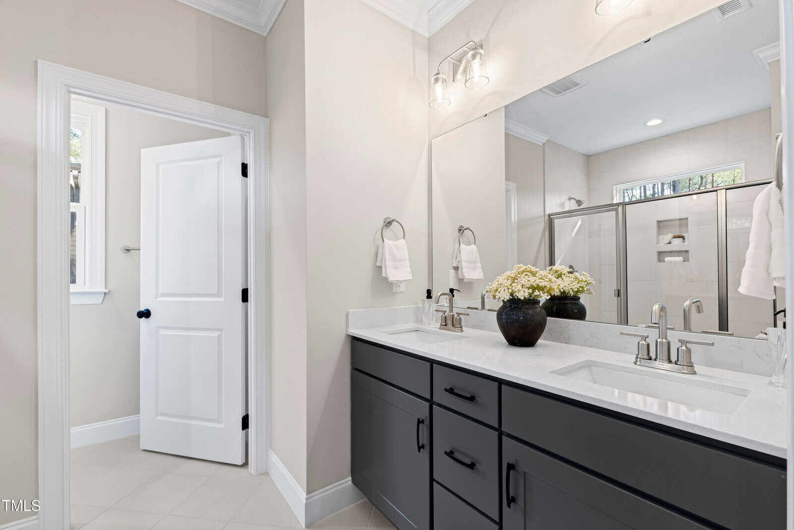 Modern bathroom with double sinks, black cabinets, large mirror, two light fixtures, and a glass-enclosed shower. White walls and countertops, a vase with flowers on the counter, and two hand towels on racks.