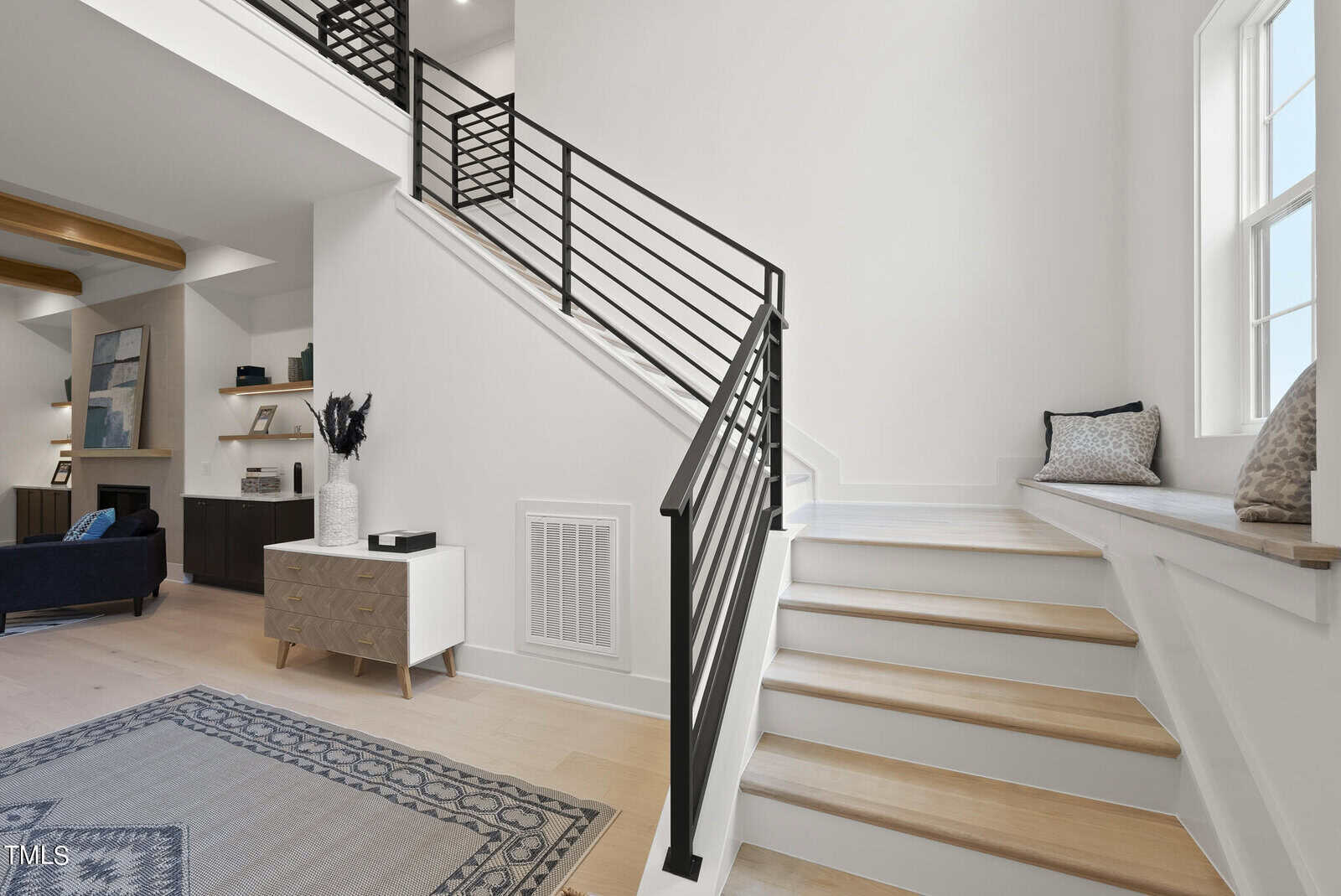 Modern, bright interior featuring a staircase with black railings, light wood steps, a window seat with cushions, a small sideboard, and a cozy living area with built-in shelving and decorative accents.