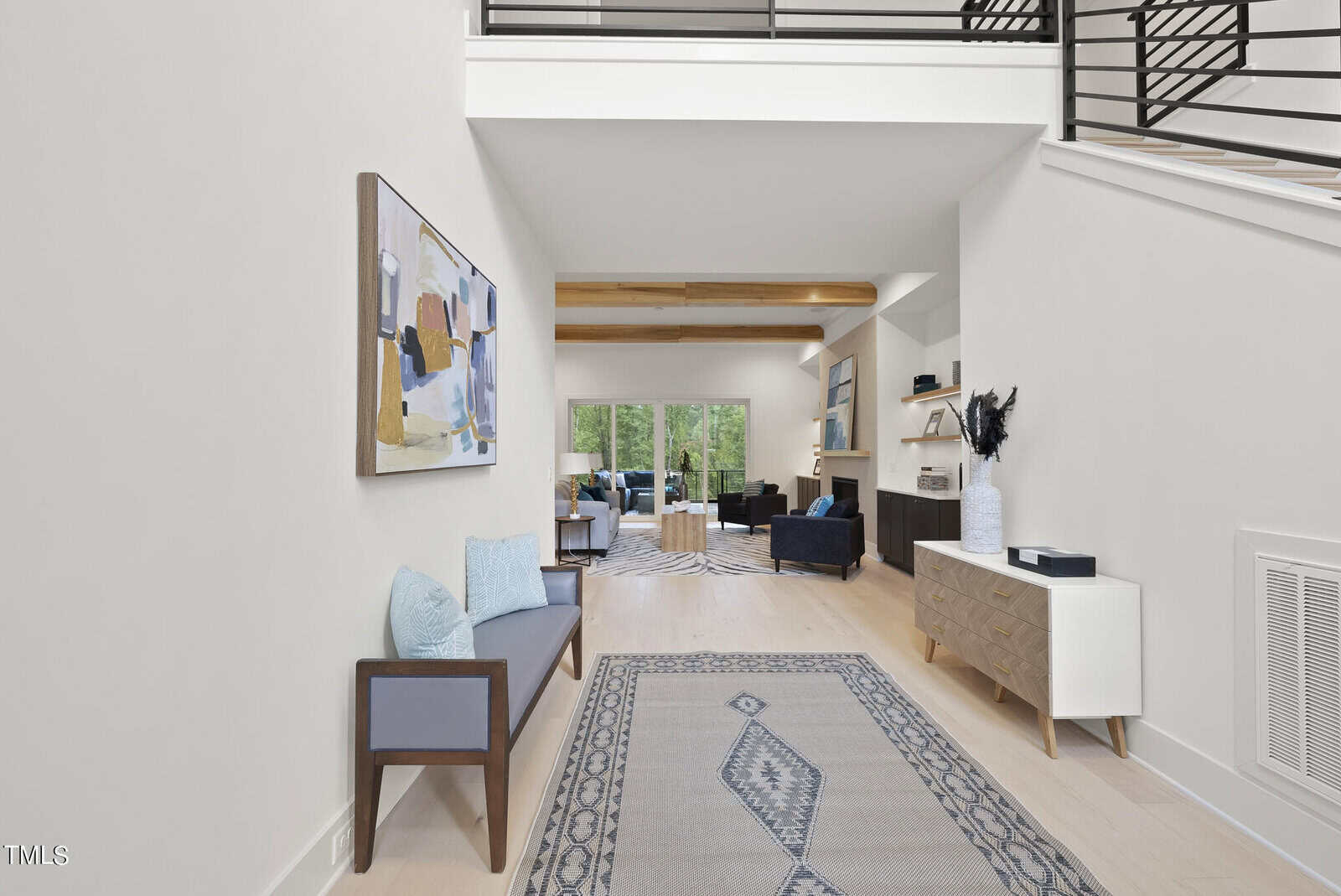 A modern, bright entryway with a patterned rug, bench with blue cushions, abstract wall art, a white sideboard, and views into a living room with large windows and wooden ceiling beams.