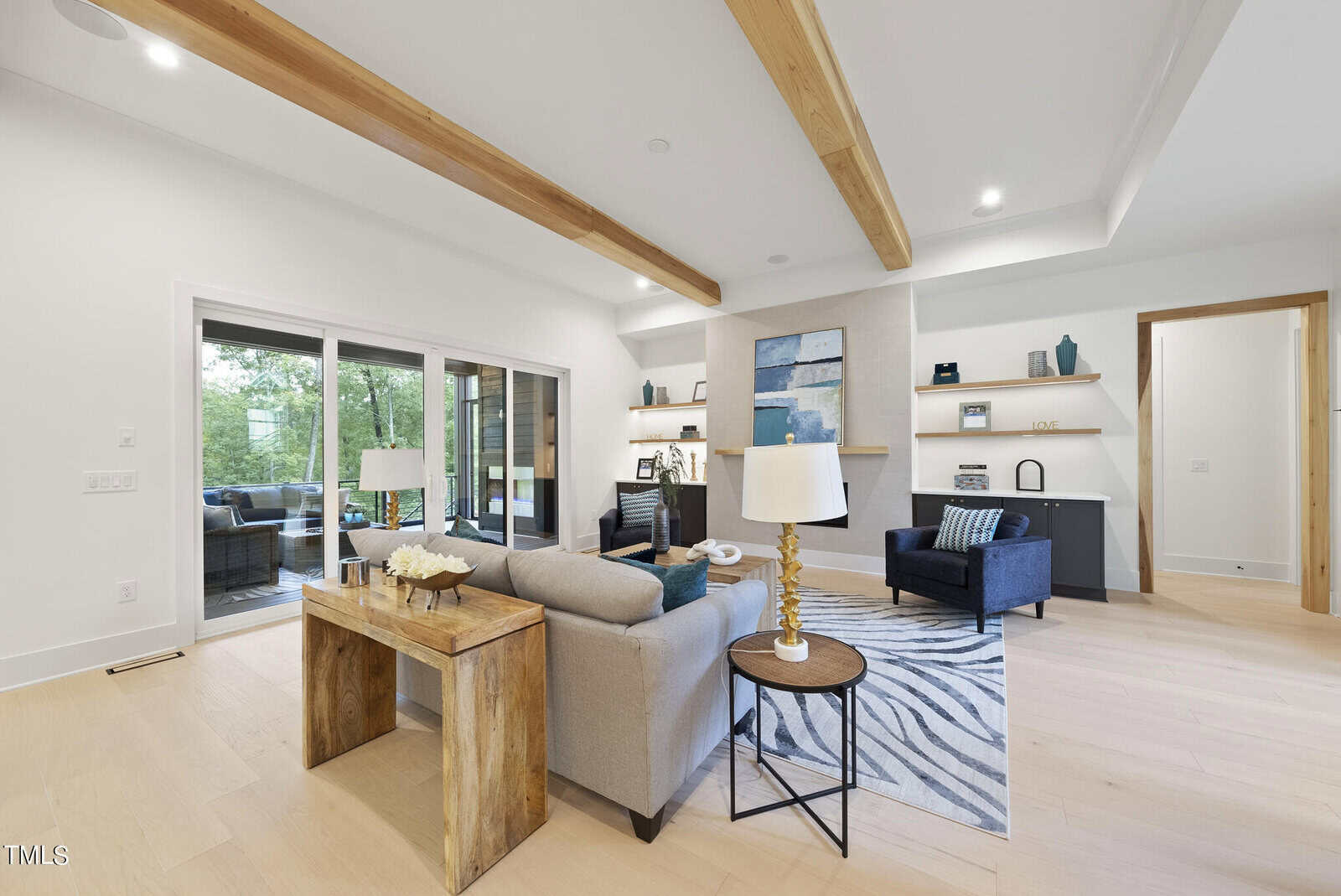 Modern living room with light wood floors, exposed ceiling beams, a gray couch, zebra rug, armchair, fireplace, built-in shelves, and sliding glass doors opening to a patio with outdoor seating and greenery visible.