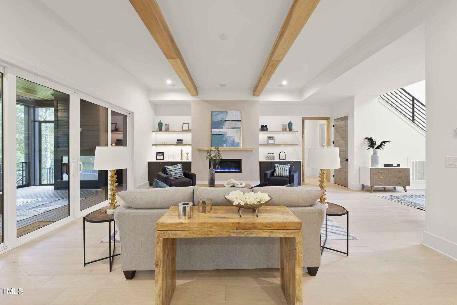 Modern living room with light wood floors, beige sofa, two lamps, wooden beams on the ceiling, a fireplace with art above it, built-in shelves, and large sliding glass doors leading to a sunroom with forest views.