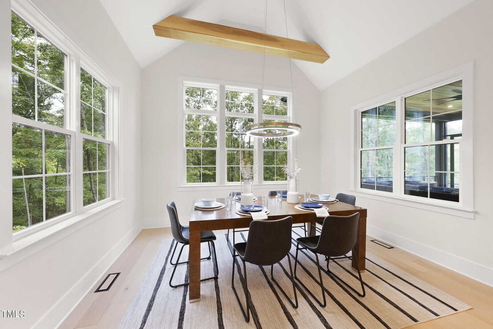 Bright dining room with large windows, a wooden table set for six, black chairs, a striped rug, modern light fixtures, and views of greenery outside. The room has light walls and hardwood floors.