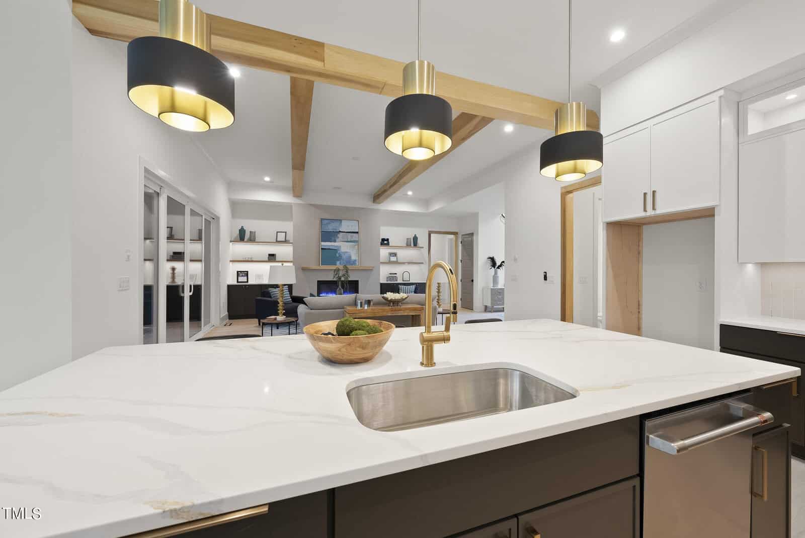 Modern kitchen with a marble island countertop, gold faucet, three black-and-gold pendant lights, and an open view into a stylish living area with built-in shelves and wood ceiling beams.