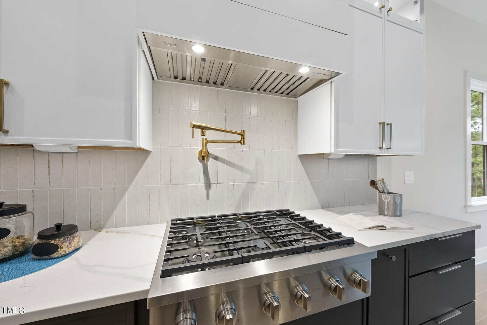 Modern kitchen stove with stainless steel gas cooktop, brass pot filler faucet, white tile backsplash, and white and black cabinetry with gold handles. Natural light comes in through a window on the right.