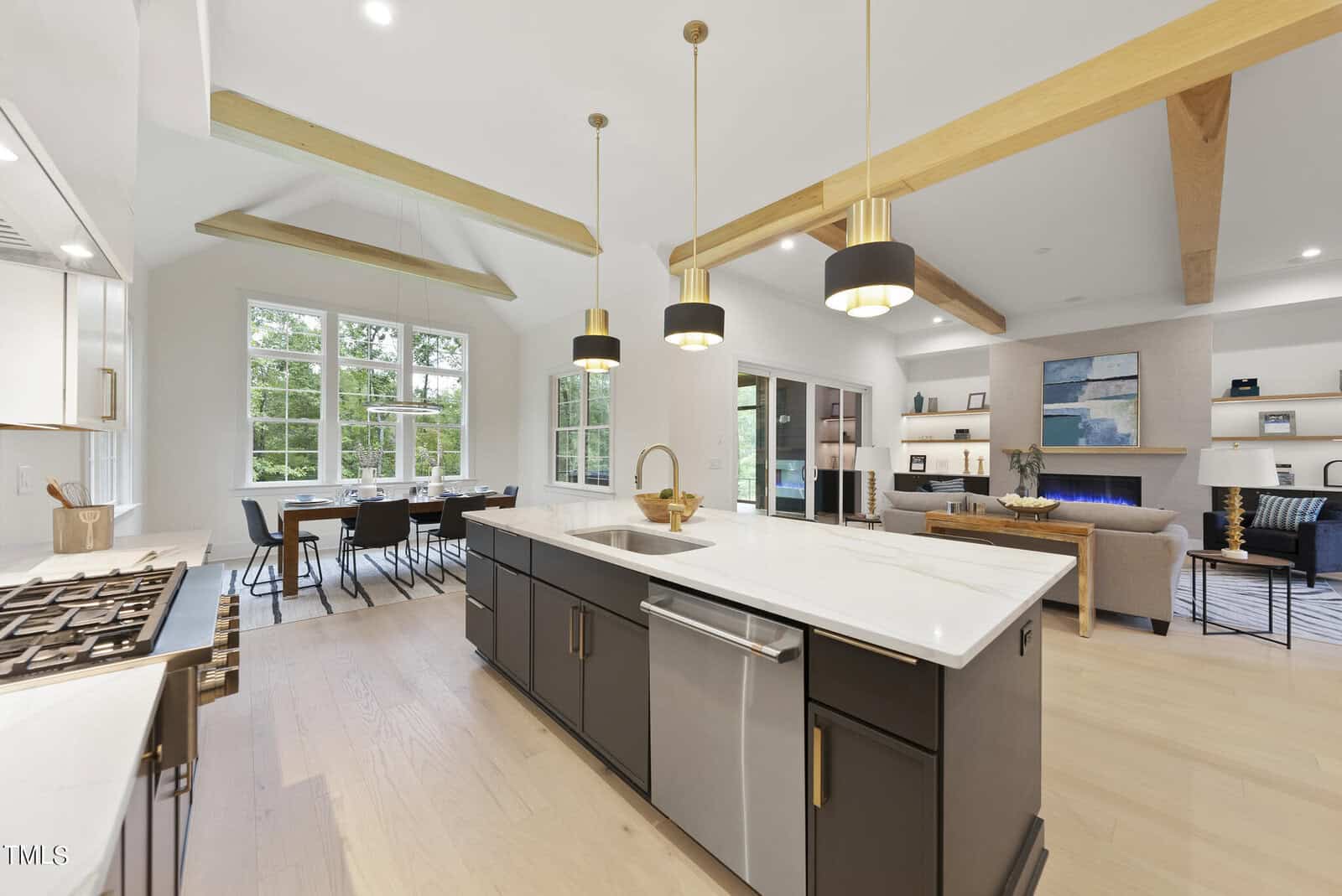 Modern open-concept kitchen and living area with wood beams, pendant lighting, large island, stainless steel appliances, dining table near windows, and a cozy living space with built-in shelves and TV.