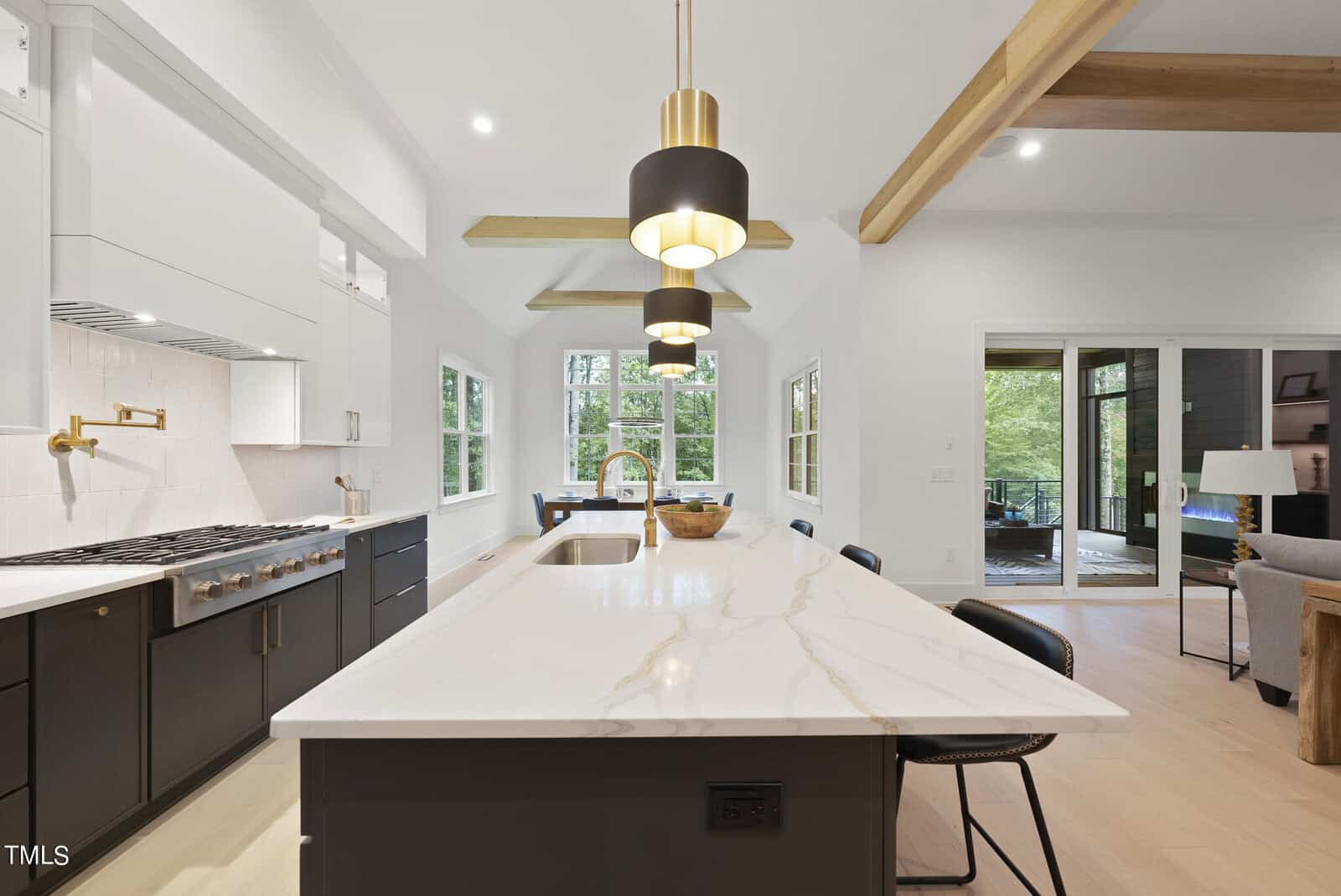 Modern kitchen with a large marble island, gold fixtures, black cabinets, pendant lights, and exposed wood ceiling beams. Large windows and sliding glass doors provide ample natural light.