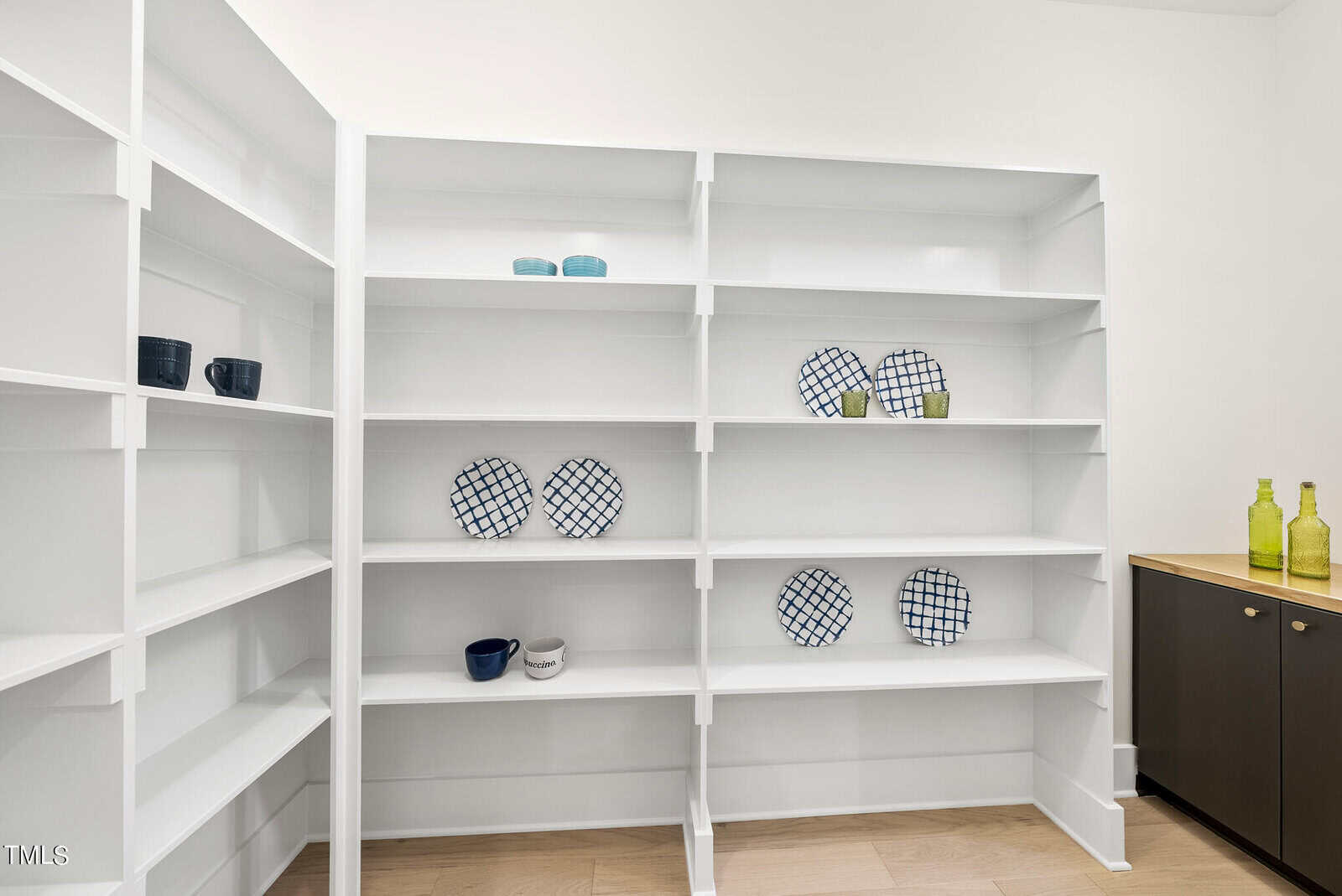 White built-in shelves in a corner display a few blue and green cups, plaid-patterned plates, and green glass bottles. The room has light wood flooring and a black cabinet with a wood countertop.