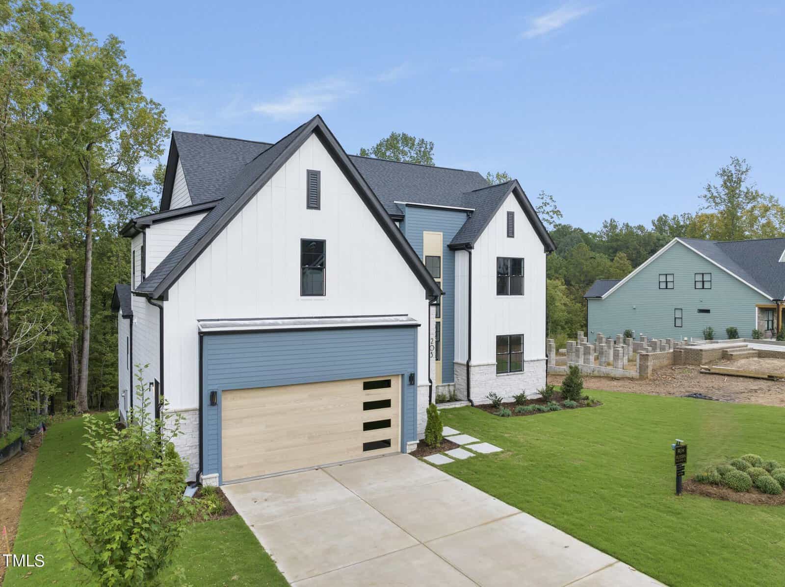 Modern two-story house with white and blue-gray exterior, black trim, and a light wood garage door. Concrete driveway, green lawn, and trees in the background; another house and construction visible nearby.