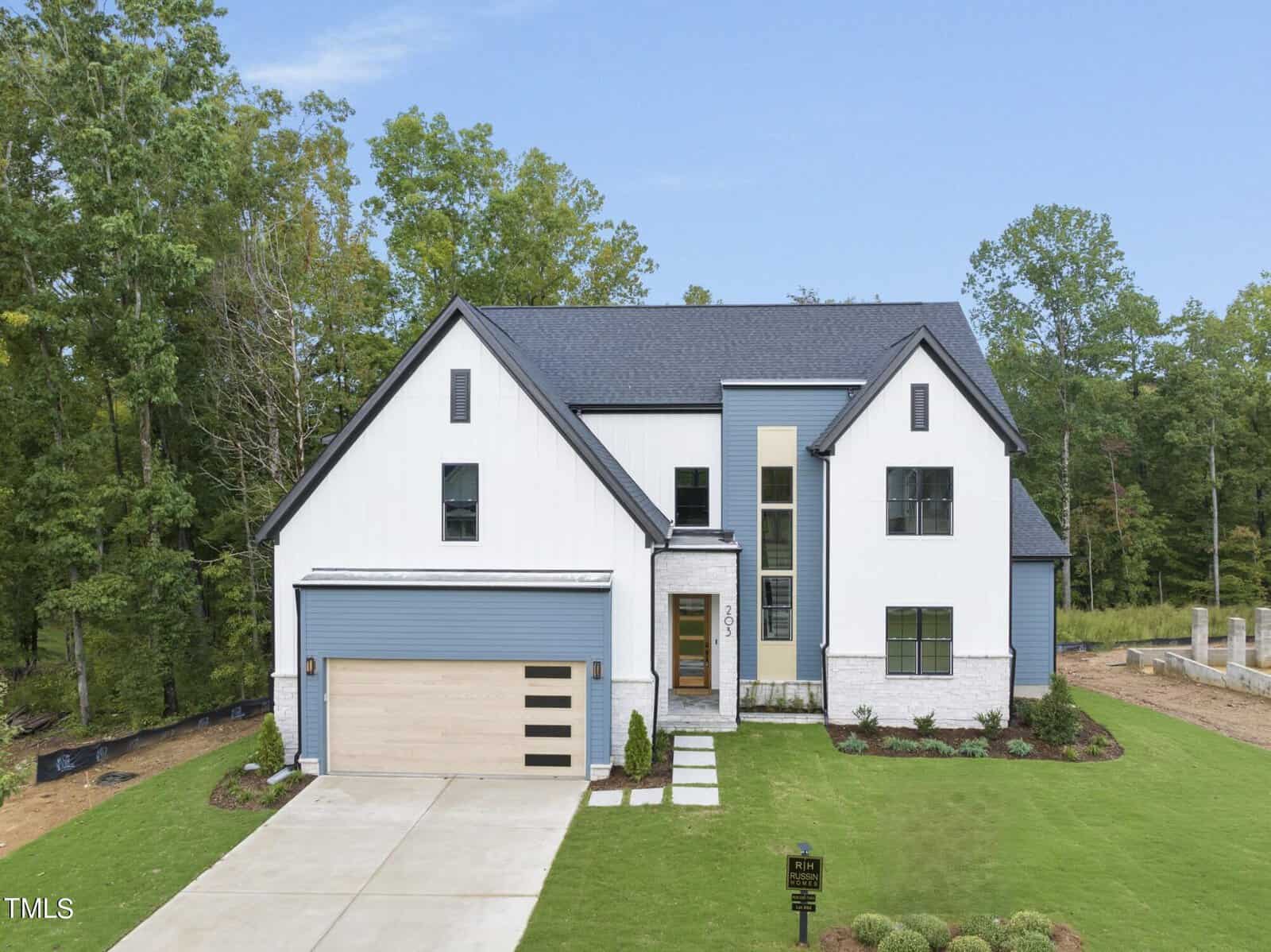 Modern two-story house with white siding, blue-gray accents, large front windows, and a double garage. The home is surrounded by green grass, neatly landscaped bushes, and bordered by tall trees.