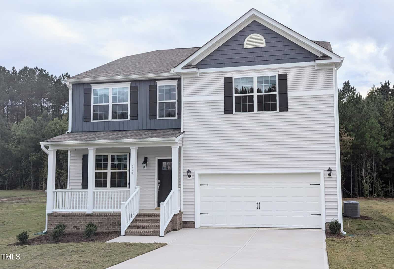 Two-story suburban house with gray and white siding, a covered front porch, black shutters, and a double garage. The house is surrounded by grass and trees in the background.