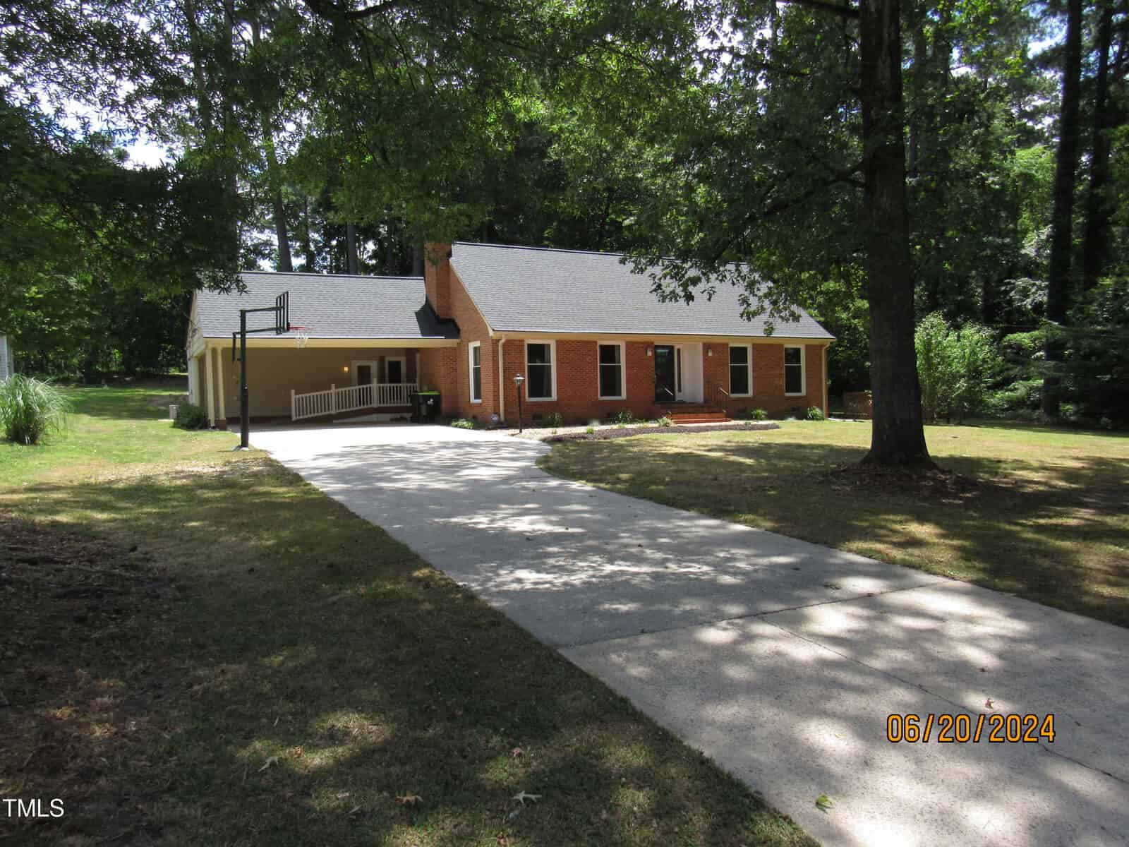 A single-story brick house with a shingled roof is partially shaded by large trees. The house features a basketball hoop beside the driveway. The lawn is mostly green, and the driveway leads to the front entrance. Date stamp reads 06/20/2024.