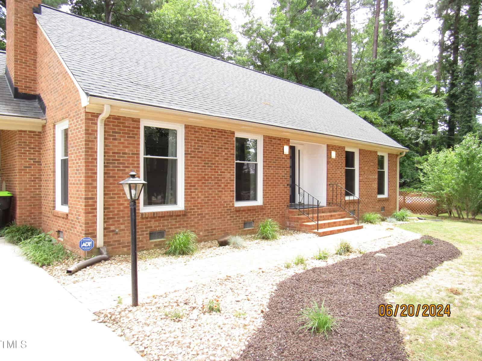 A brick house with a sloped roof, white-framed windows, and a central white door. There's a gravel path leading to the entrance, surrounded by a lawn and trees. A lamppost is near the walkway. Date-stamped as June 20, 2024.