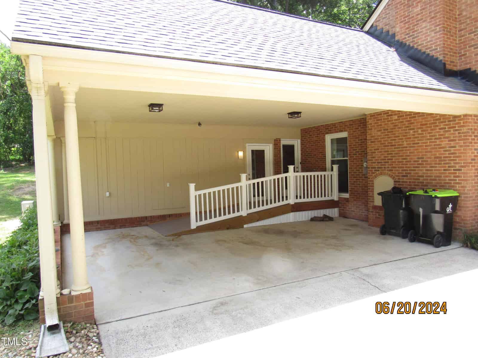 A covered carport with a sloped ramp leading to a side entrance of a brick house. There are three trash bins nearby and it is surrounded by trees. The carport has a shingled roof and two supporting columns. Date stamp in the corner reads 06/20/2024.