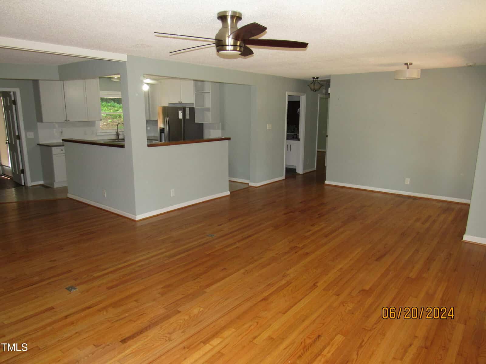 Spacious living area with wooden floors, light gray walls, and a ceiling fan. There's an open kitchen with white cabinets and stainless steel appliances. The room is well-lit, with large windows on the left, and a chandelier in the distance.