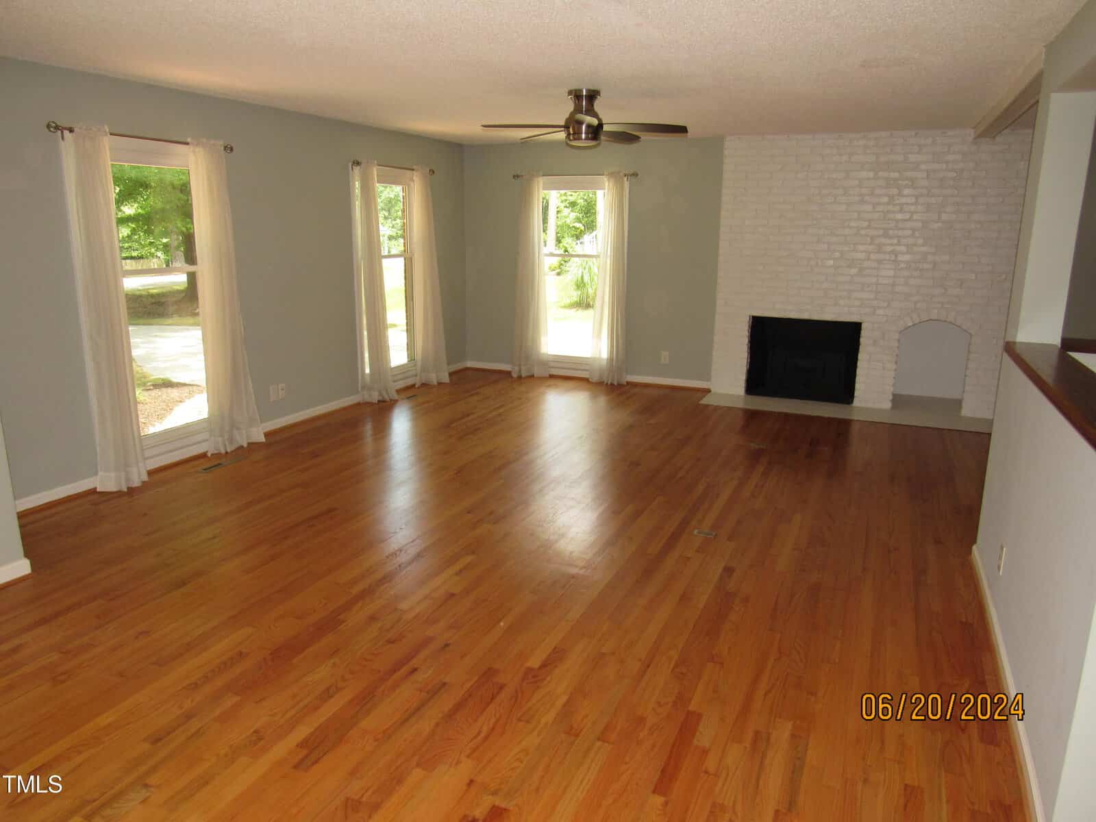 A spacious living room with wooden floors, light blue walls, and white curtains. Two large windows let in natural light. There's a ceiling fan and a white brick fireplace on the right wall.