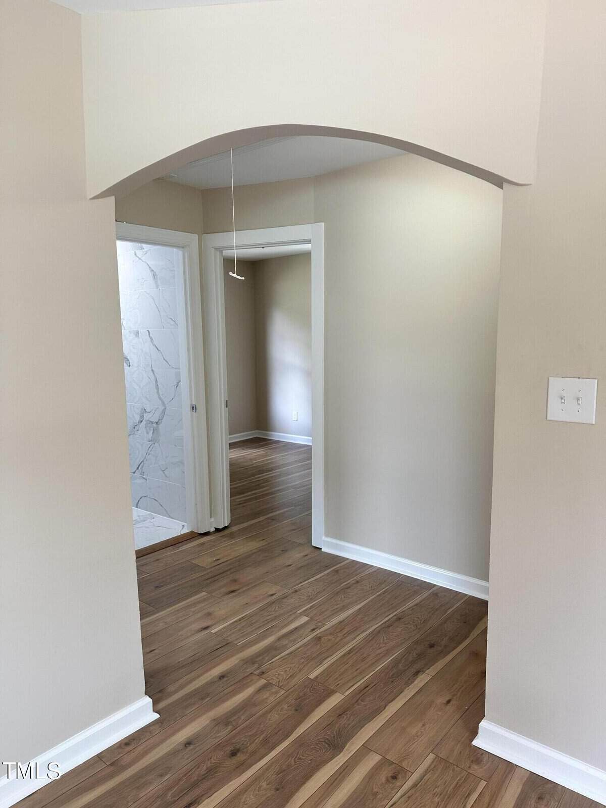A hallway with light beige walls and wooden floors. There is an arched doorway leading to a room in the background. On the left, a partially open door shows a tiled wall in a bathroom. A light switch is on the right wall.