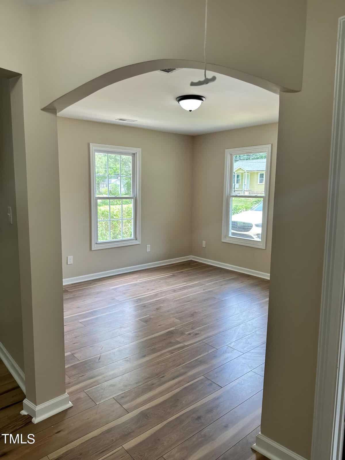 A well-lit empty room with light-colored walls, wooden flooring, and two large windows allowing natural light to flood in. The room features an arched opening, a recessed ceiling light, and a view of green trees and a yellow house through the windows.