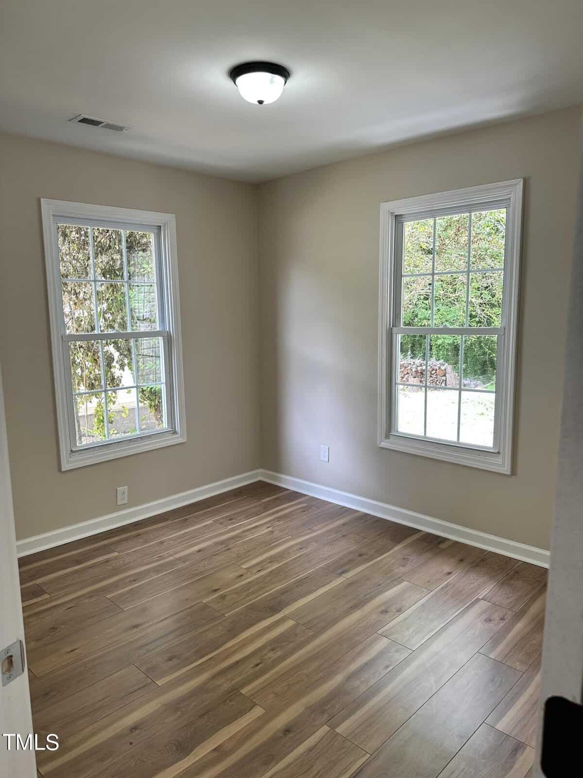 A small empty room features two large windows with white frames, beige walls, and wood-patterned laminate flooring. A single round ceiling light fixture illuminates the space, and outside, trees and greenery are visible through the windows.