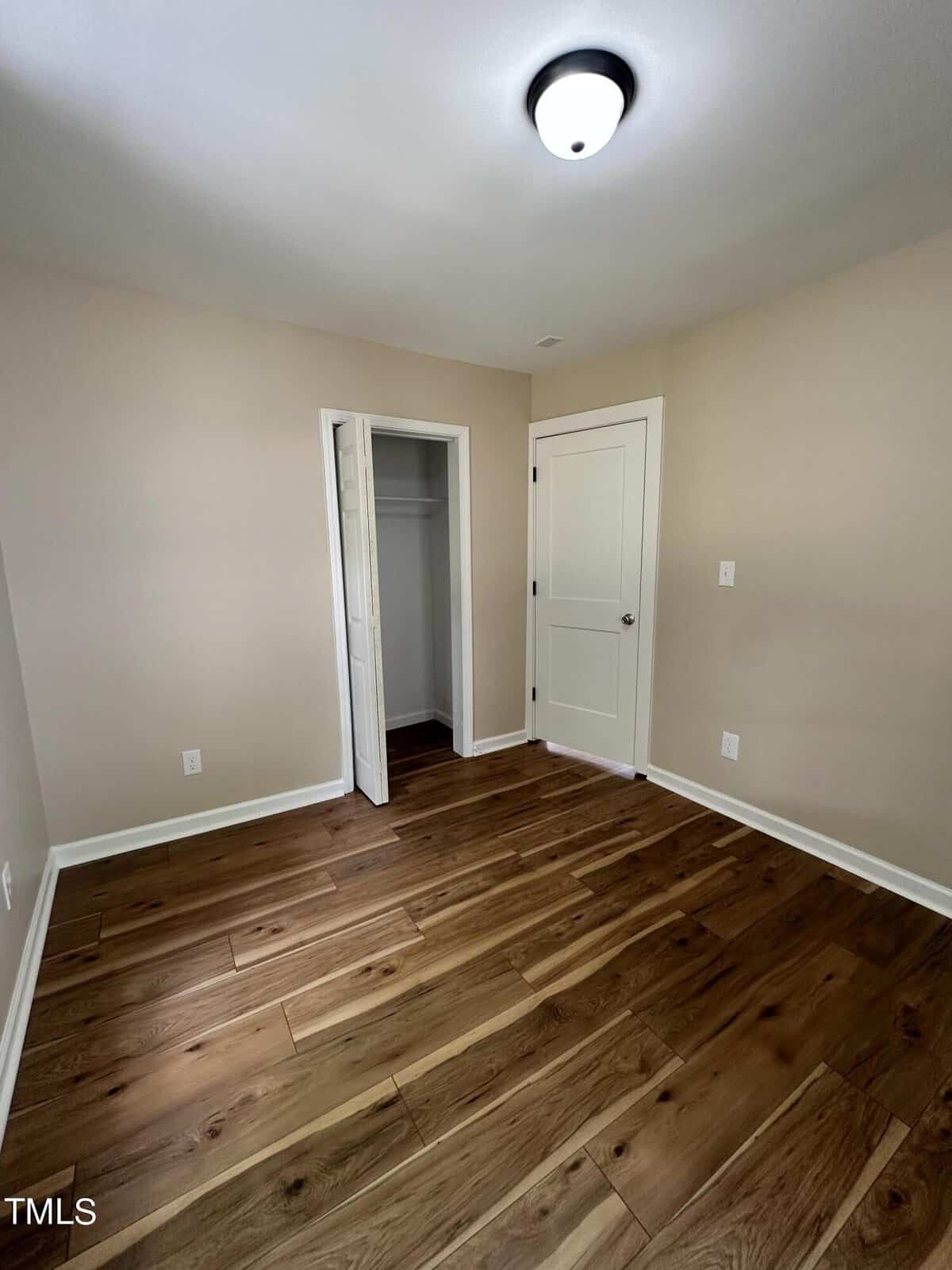 An empty room with light beige walls, a ceiling light, and dark hardwood floors. It features a small closet with an open door and a closed white interior door. The room is well-lit by natural light.