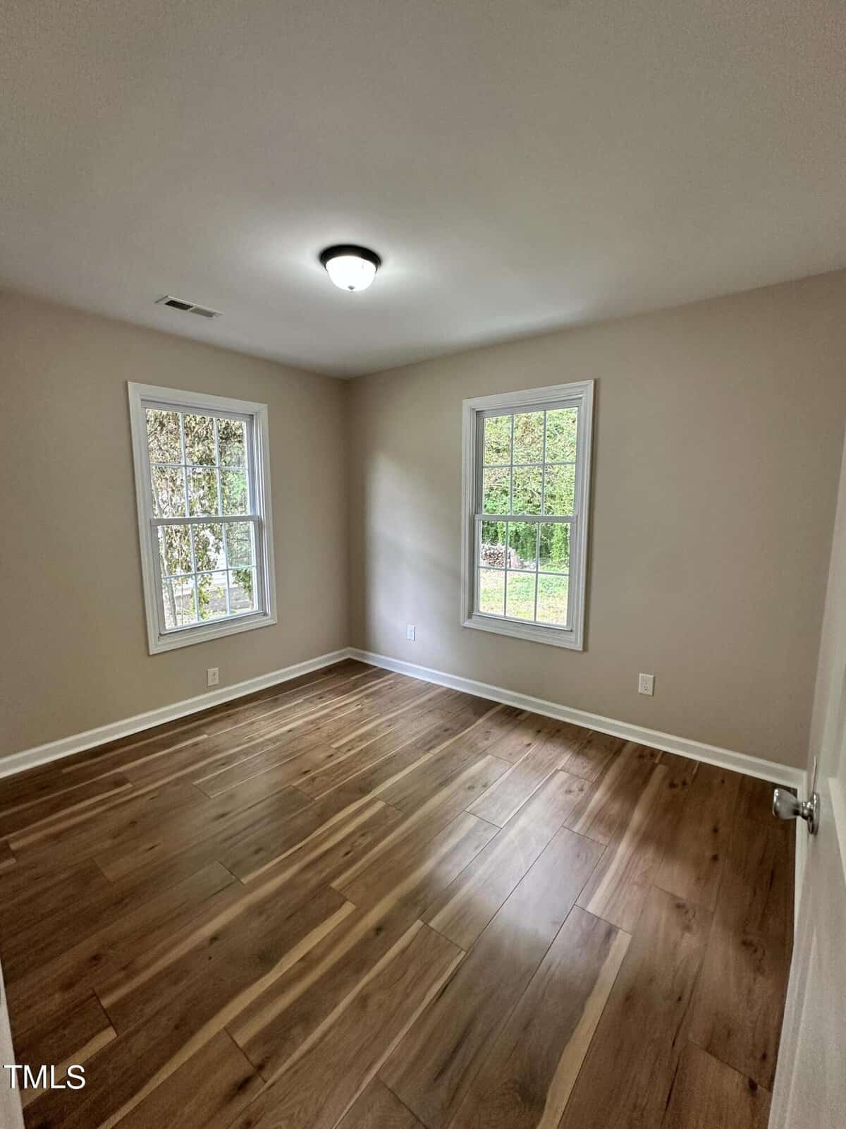 A small, empty room with light-colored walls and wood flooring. Two large windows allow natural light to enter, offering a view of greenery outside. A ceiling light fixture is centered on the white ceiling. The room has a minimalist and clean design.