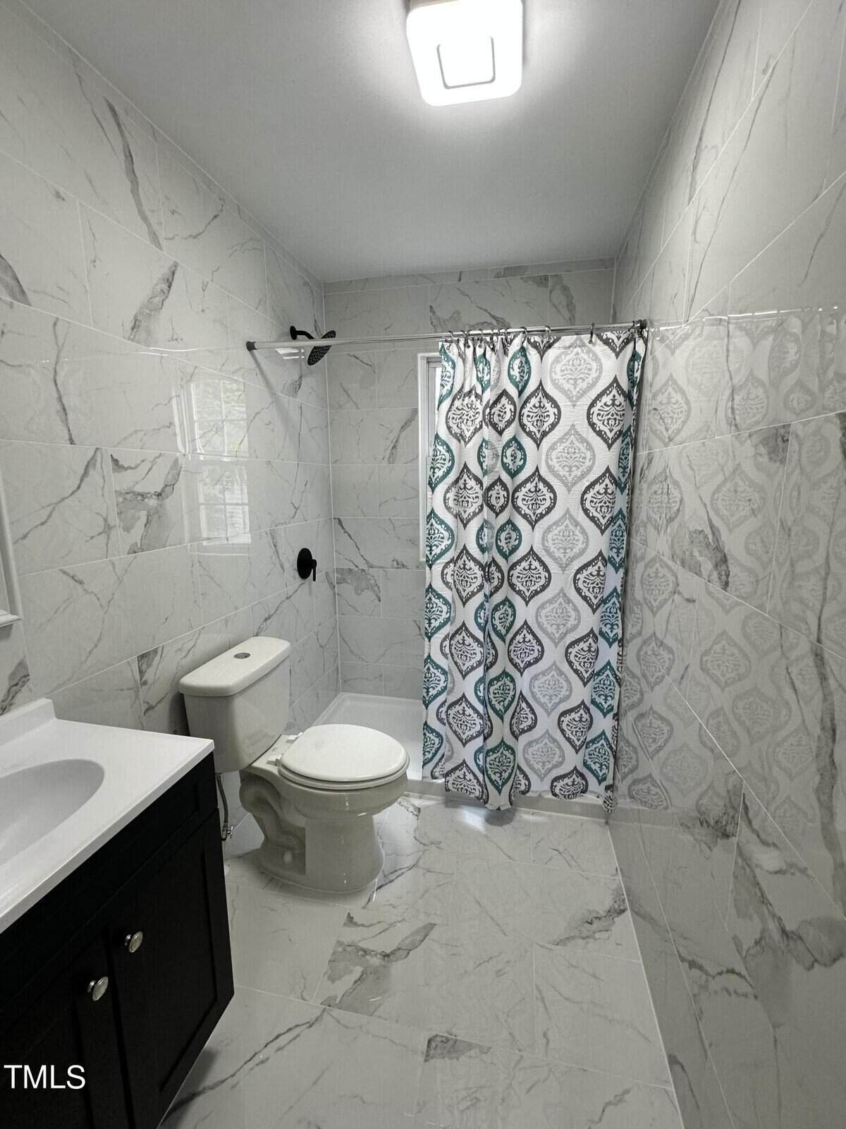 A bathroom with white marble tile walls and floors. It features a toilet, a dark wood vanity with a white countertop, and a shower with a patterned curtain in blue, white, and gray tones. A rectangular ceiling light fixture illuminates the space.
